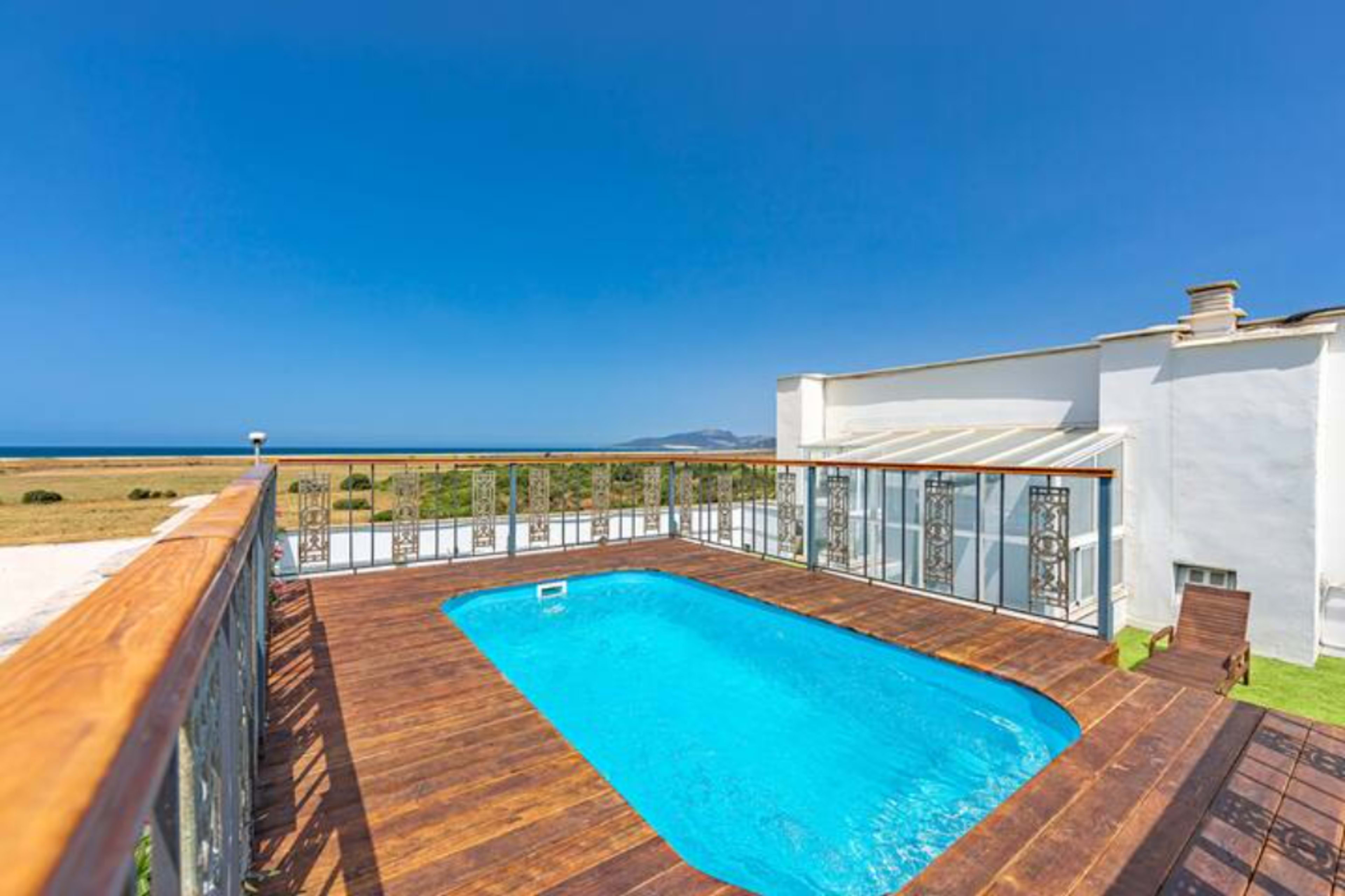 A modern swimming pool surrounded by wooden decking, adjacent to a white building with large glass windows, overlooking a clear blue sea and a grassy landscape.