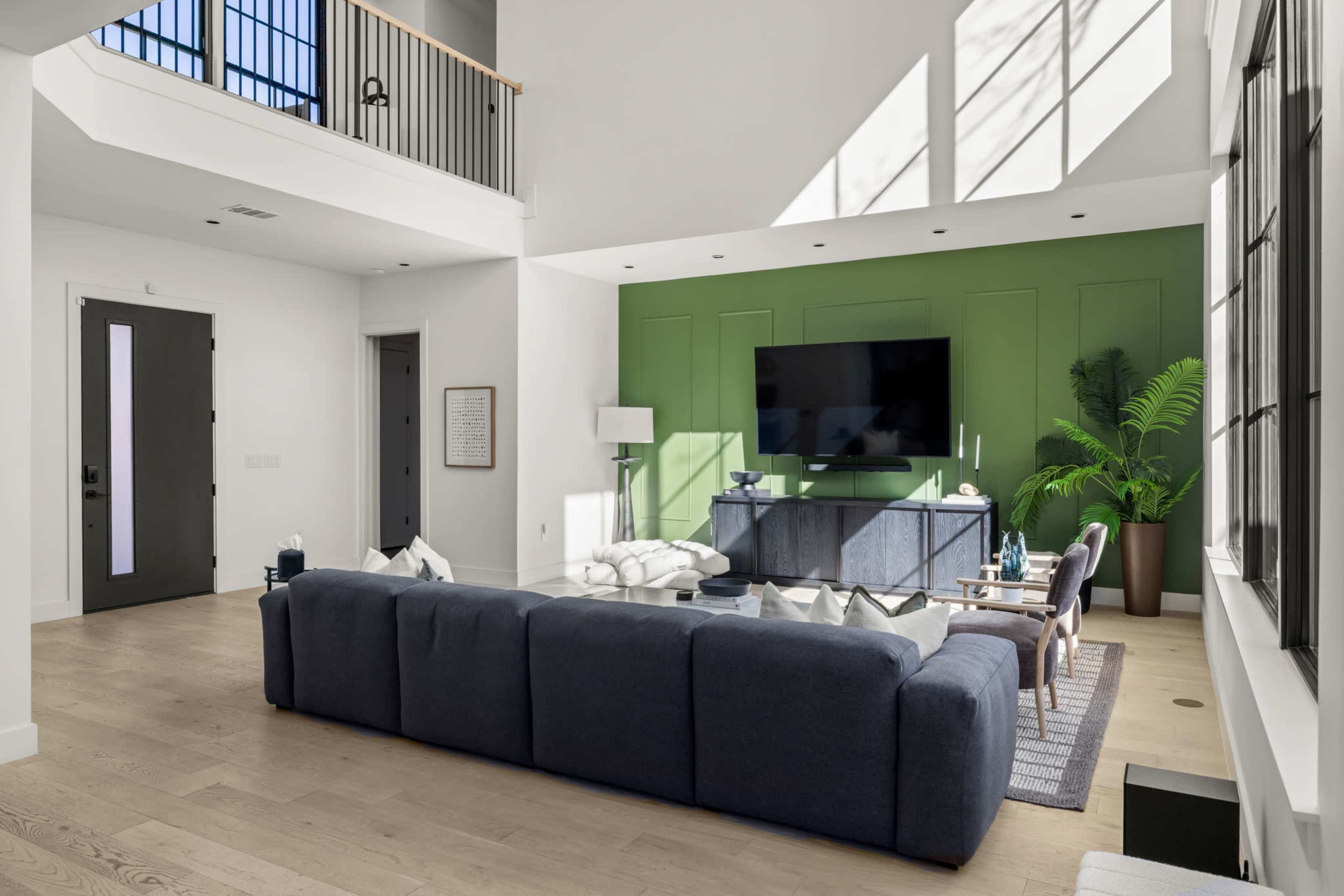 A spacious living room features a dark blue sectional sofa facing a large TV mounted on a green accent wall, with natural light streaming in through tall windows.