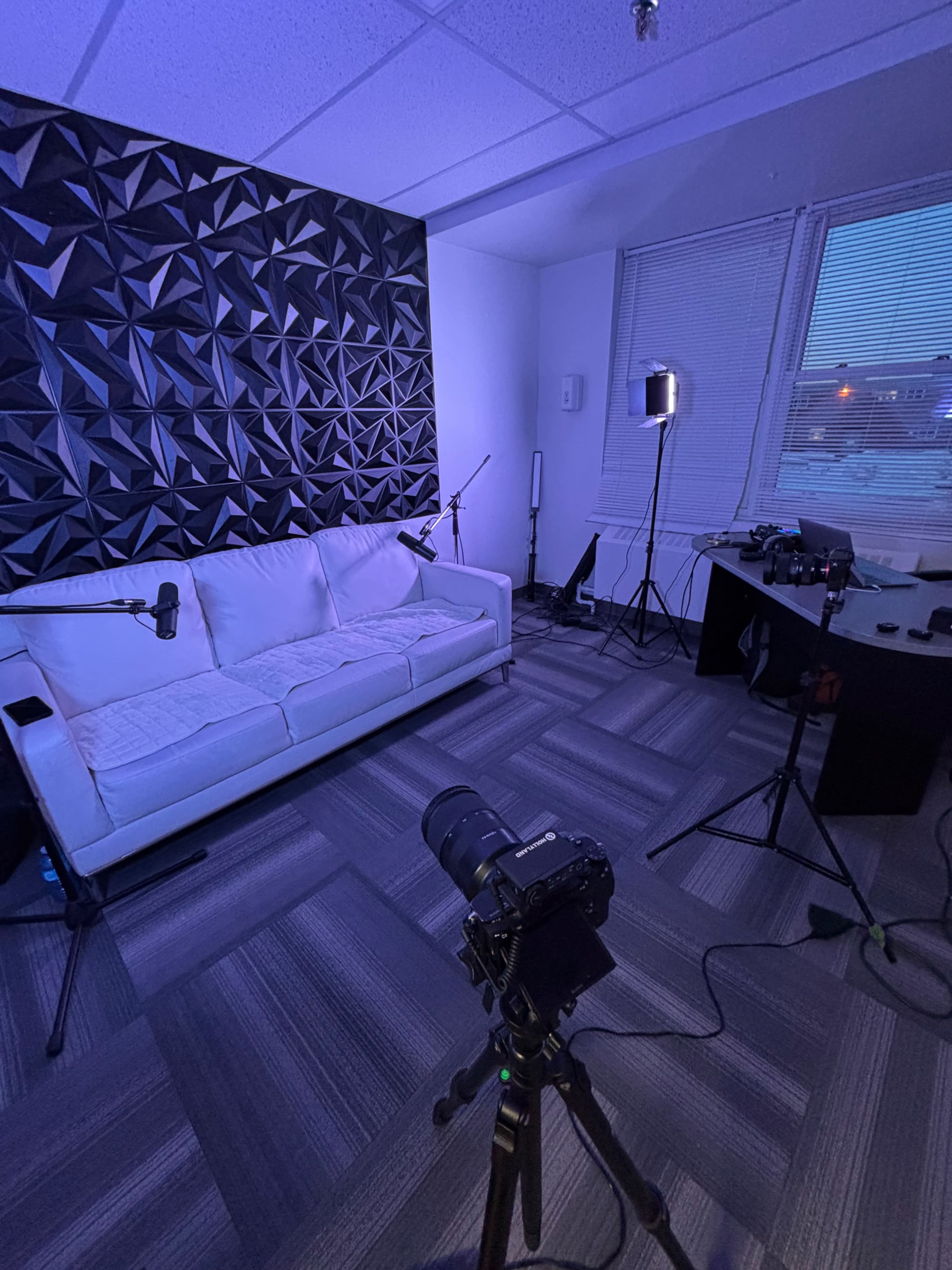 The image shows a modern studio setup featuring a white couch, a camera on a tripod, and various lighting equipment arranged in a well-lit room with large windows.