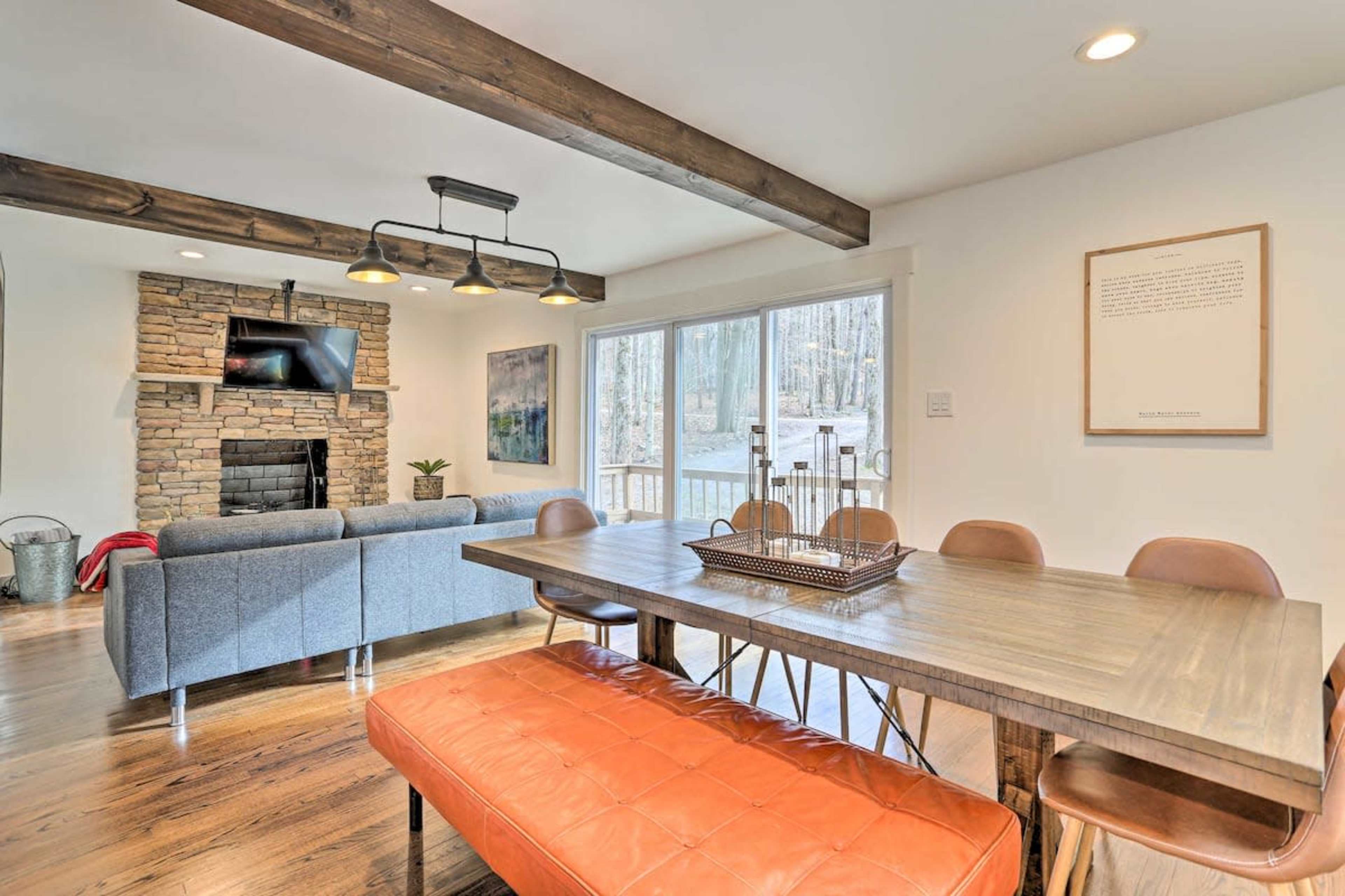 A dining area with a wooden table and orange bench faces a stone fireplace and a large window that offers a view of a wooded area.