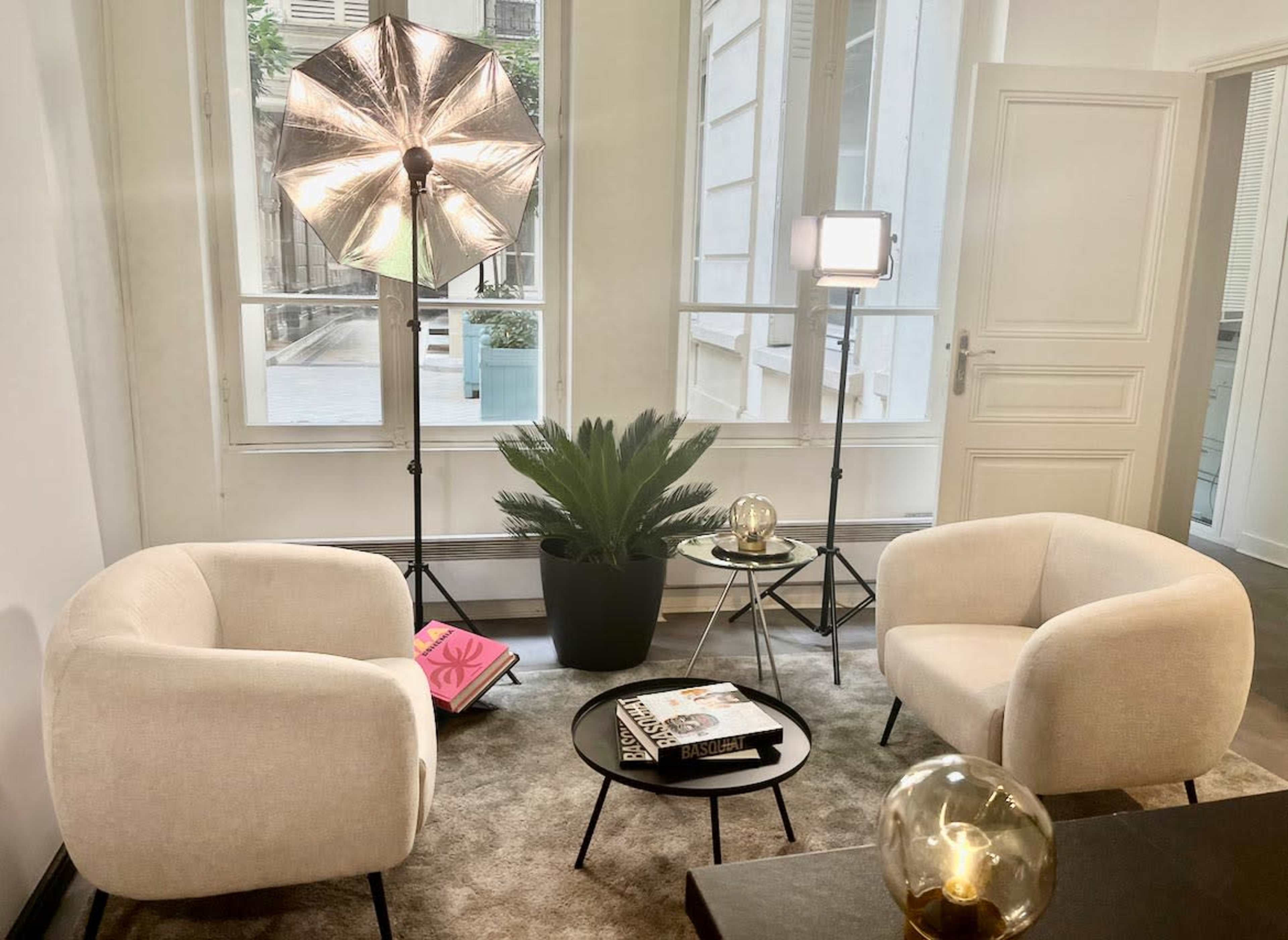 The image shows a modern, well-lit interior space featuring two light-colored chairs, a small black table, and a potted plant, with photography equipment set up nearby.