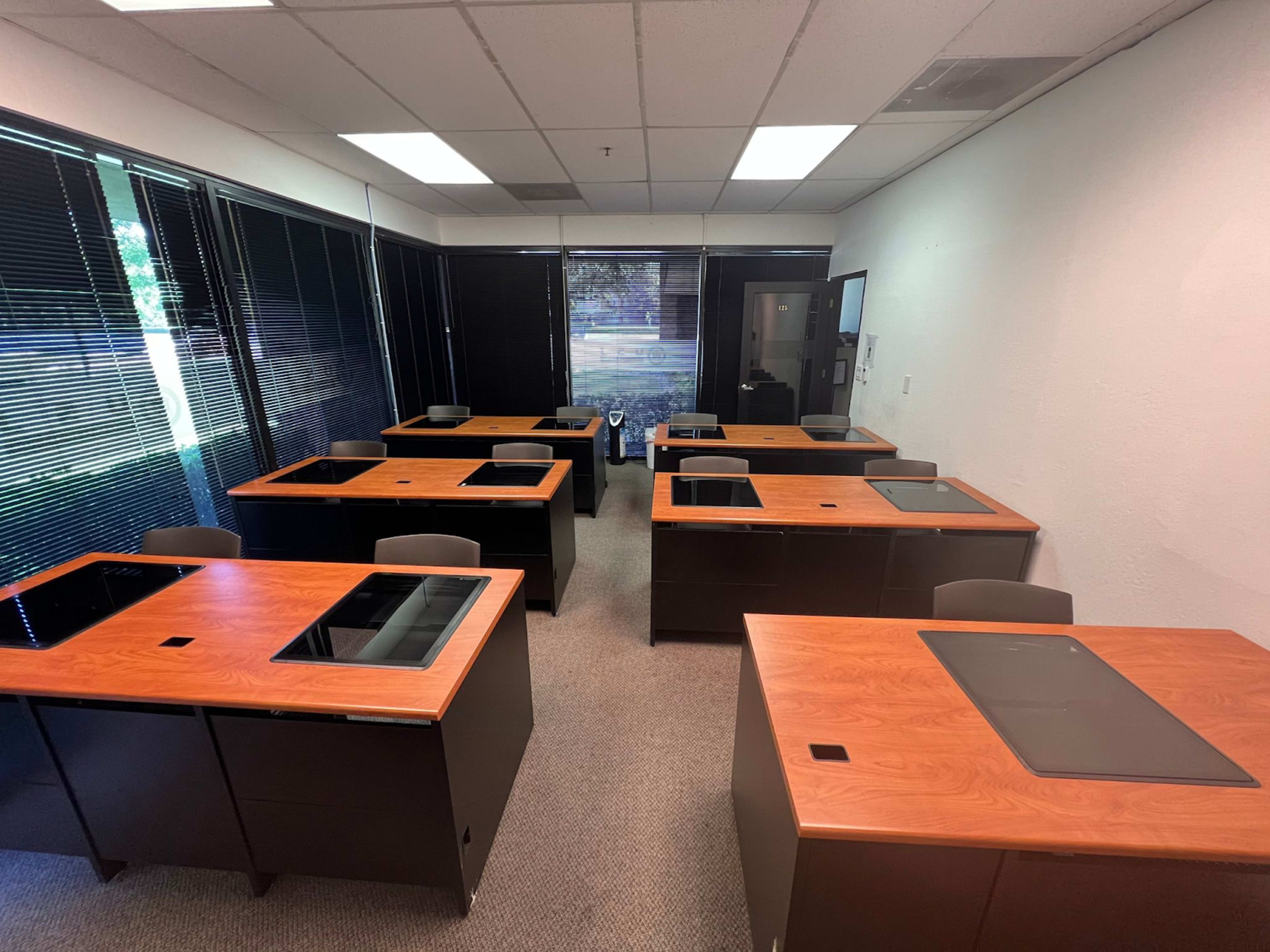 A spacious office room with multiple desks arranged in rows, each featuring a wooden surface and a chair.