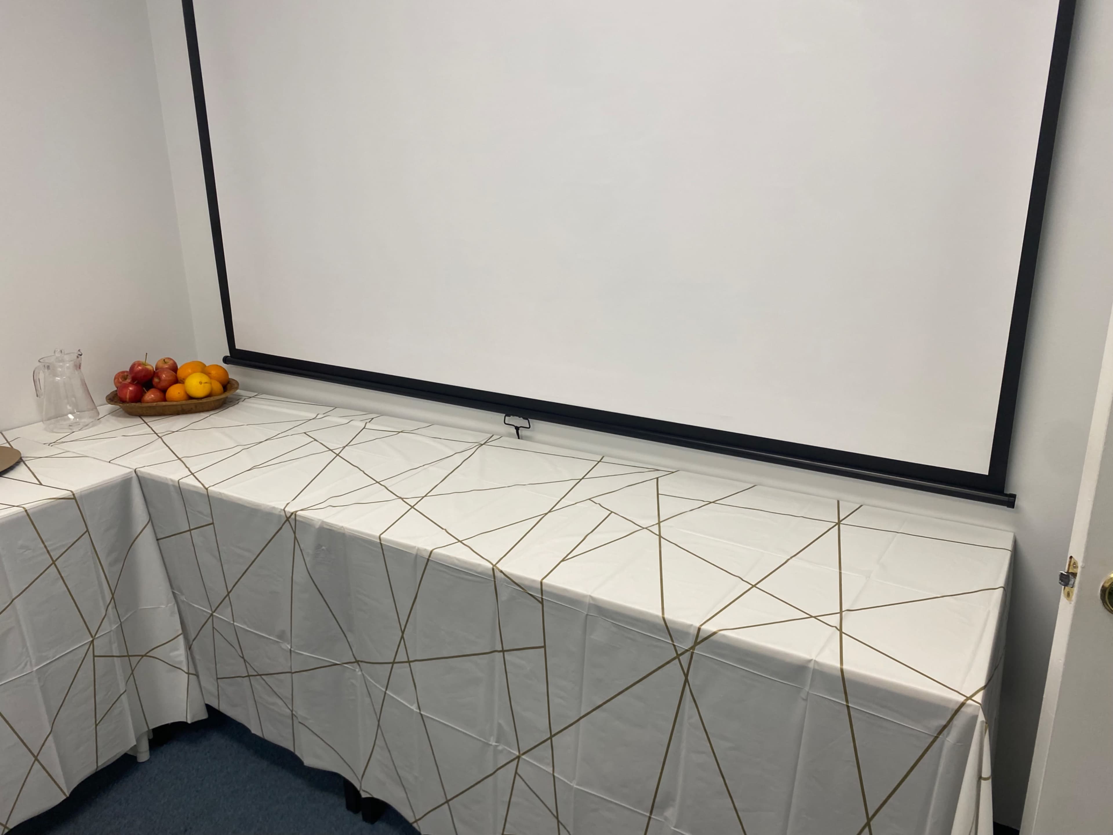 A table covered with a white tablecloth featuring a geometric gold design is set up in front of a blank projection screen, with a bowl of fruit displayed on the left.