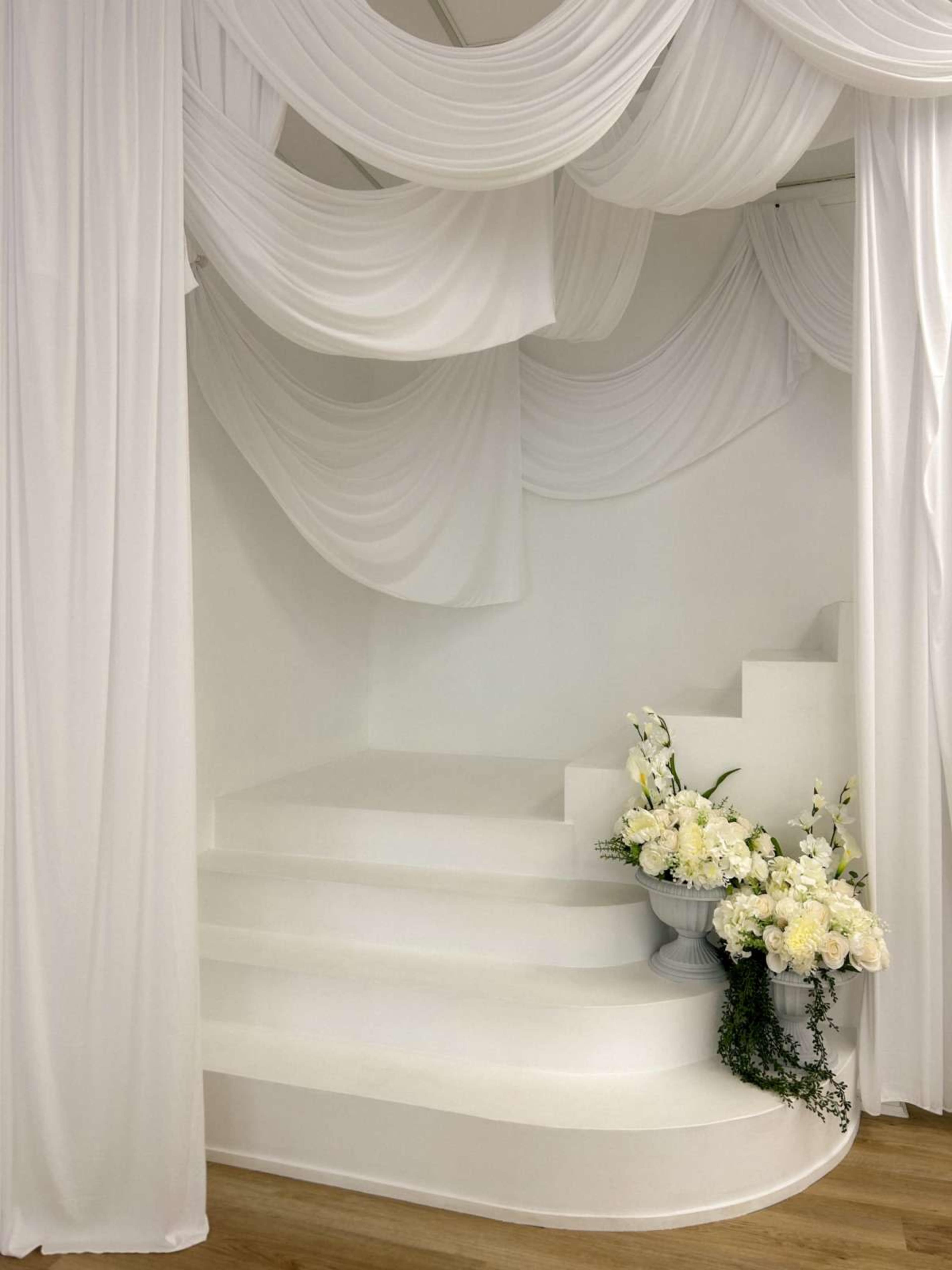 The image shows a white draped interior with a set of steps leading upward, accented by a floral arrangement in a vase on the side.