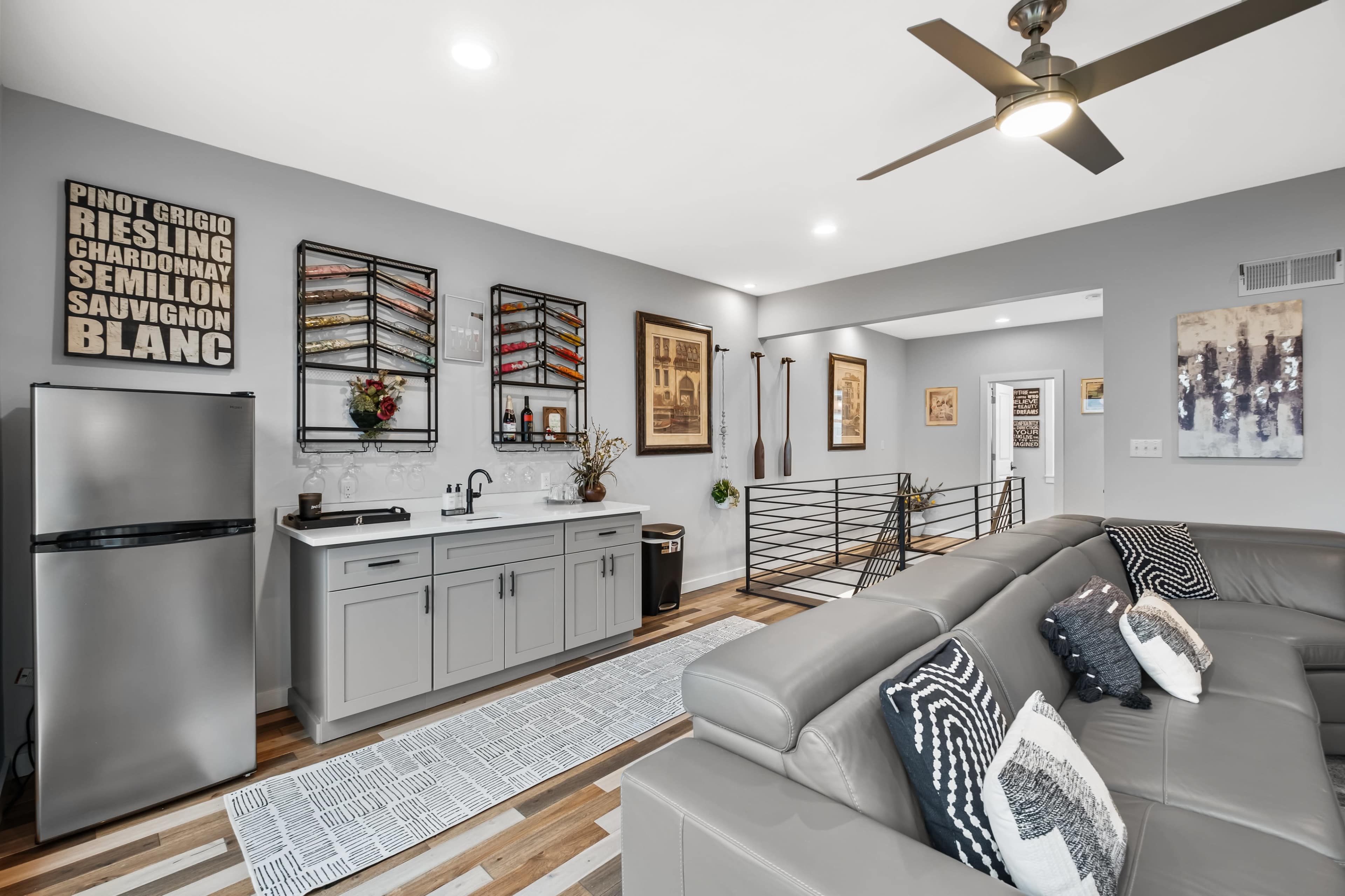 The image shows a modern living space featuring a gray sofa, a small kitchen area with a mini-fridge, and decorative wall art in a well-lit room.