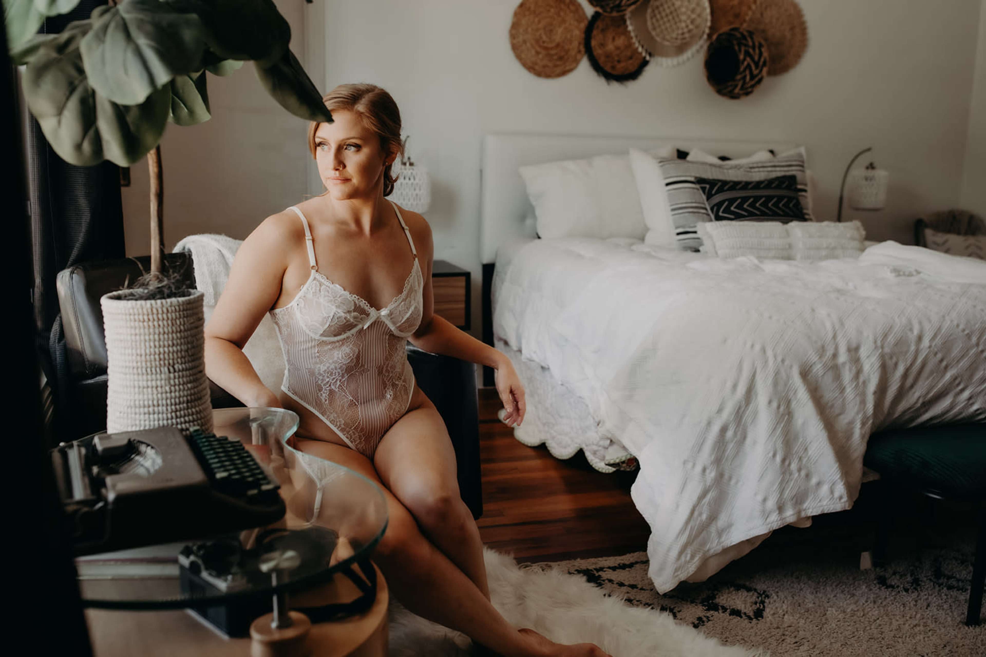 A woman sits on a chair in a bedroom, wearing white lingerie, with a bed and decorative elements visible in the background.