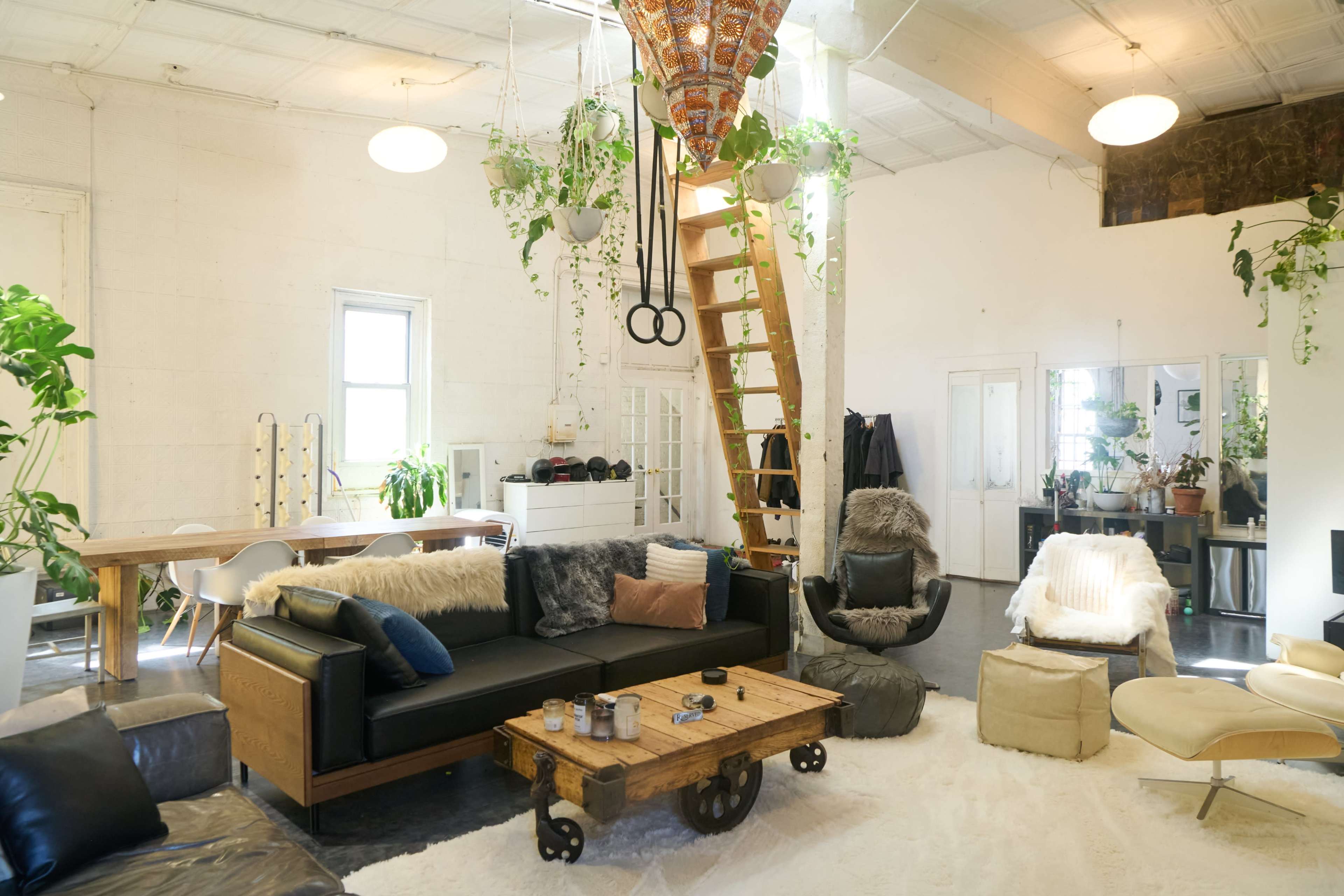 The image shows a spacious, bright living area with a mix of modern furniture, plants, and a wooden staircase leading to a lofted area.