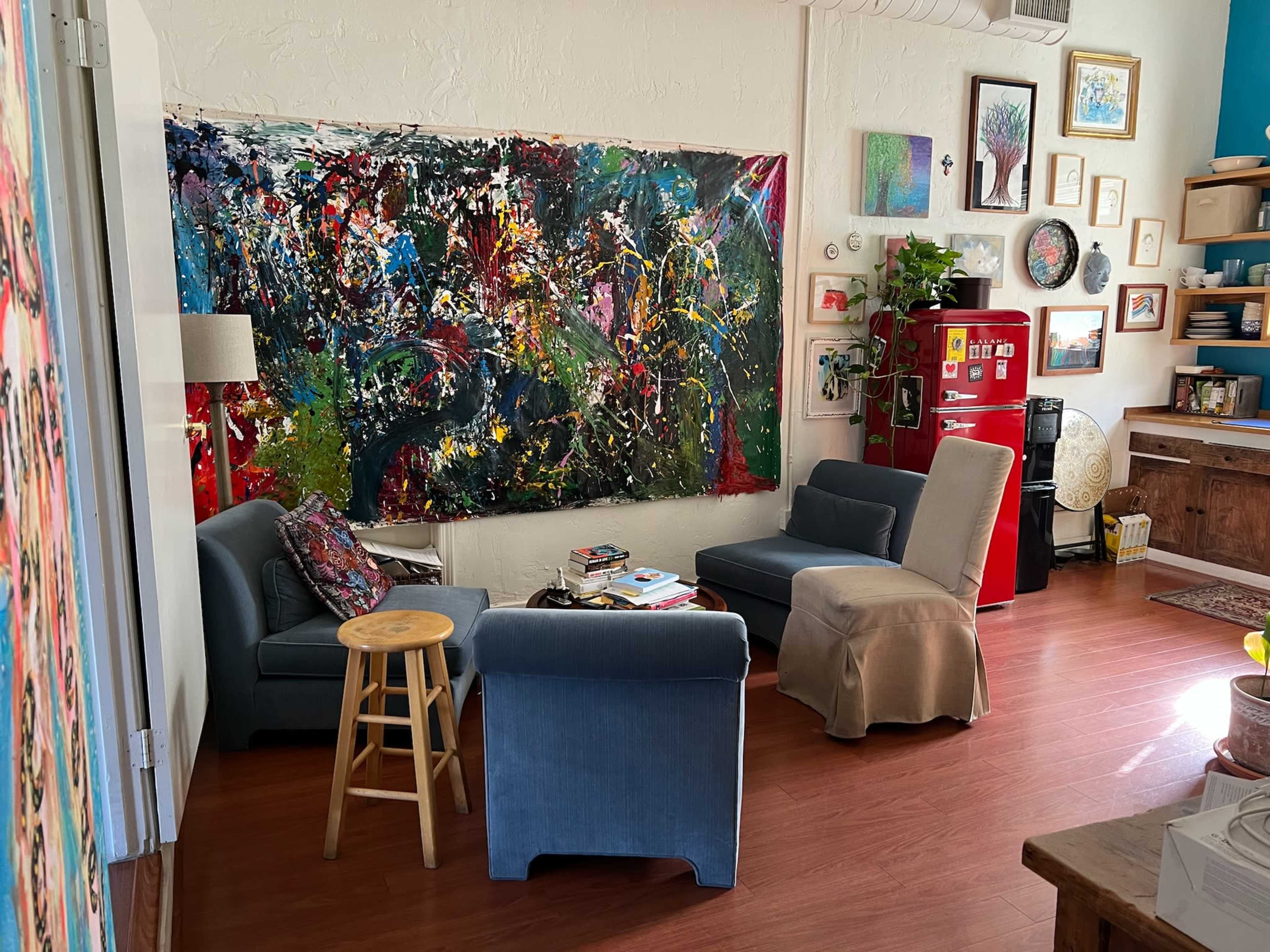 A cozy living area with a colorful abstract painting on the wall, two upholstered chairs, a stool, and a small table, all surrounded by various decorations and furniture.