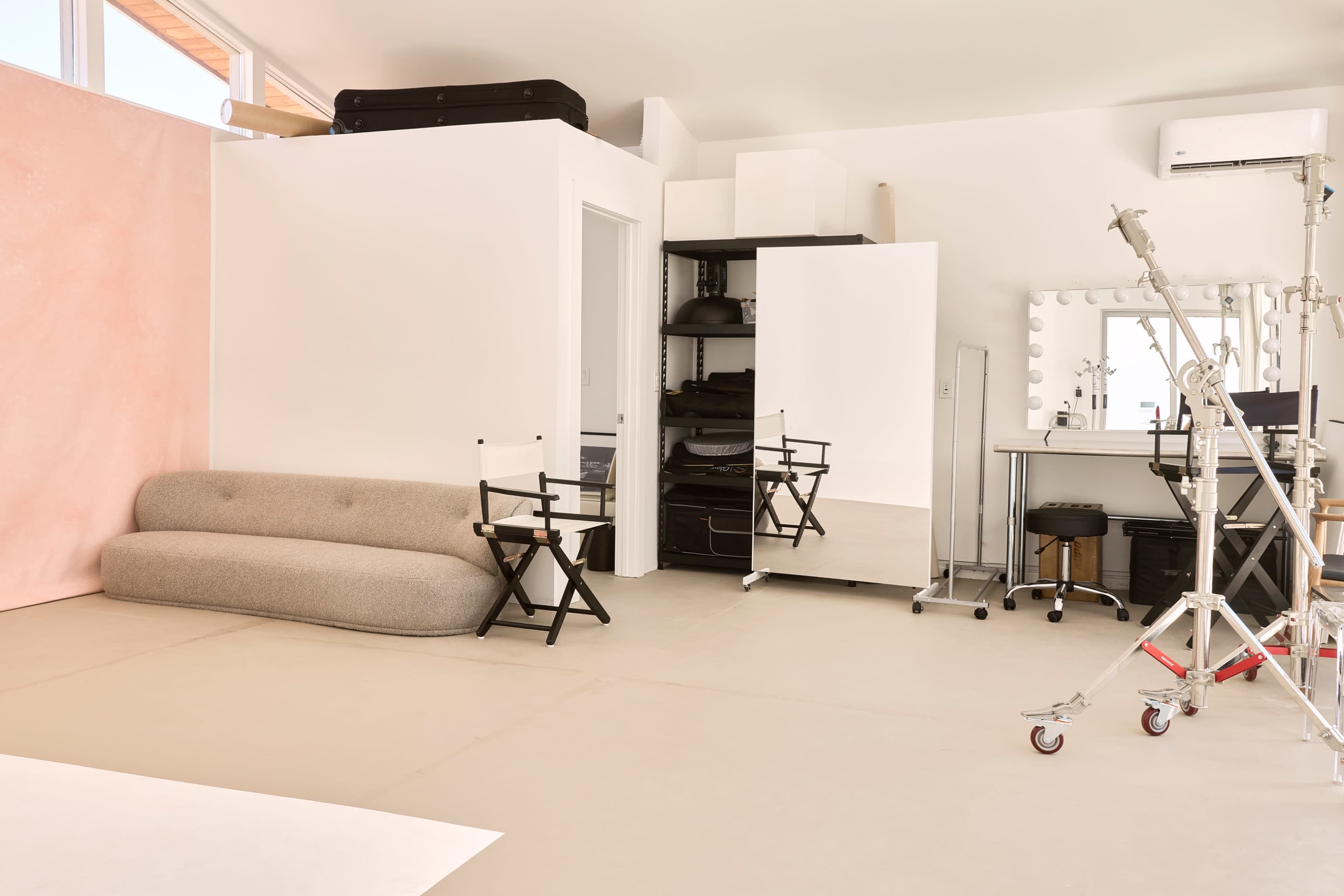 The image shows a spacious, modern studio featuring a pink backdrop, a mirrored wardrobe, and various furniture pieces including a sofa and director's chairs.