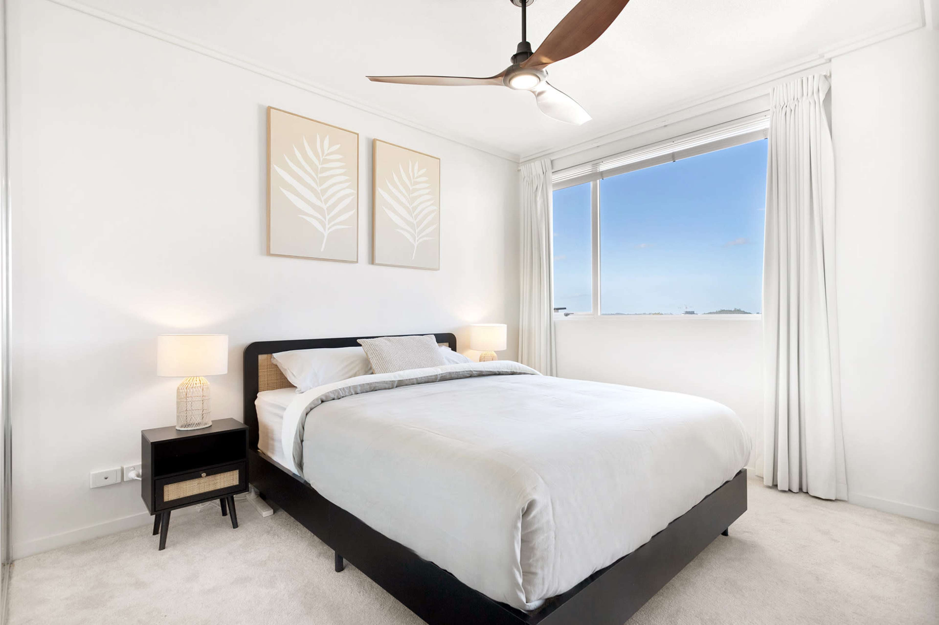 A well-lit bedroom features a large bed, nightstands with lamps, and framed plant artworks on the wall.