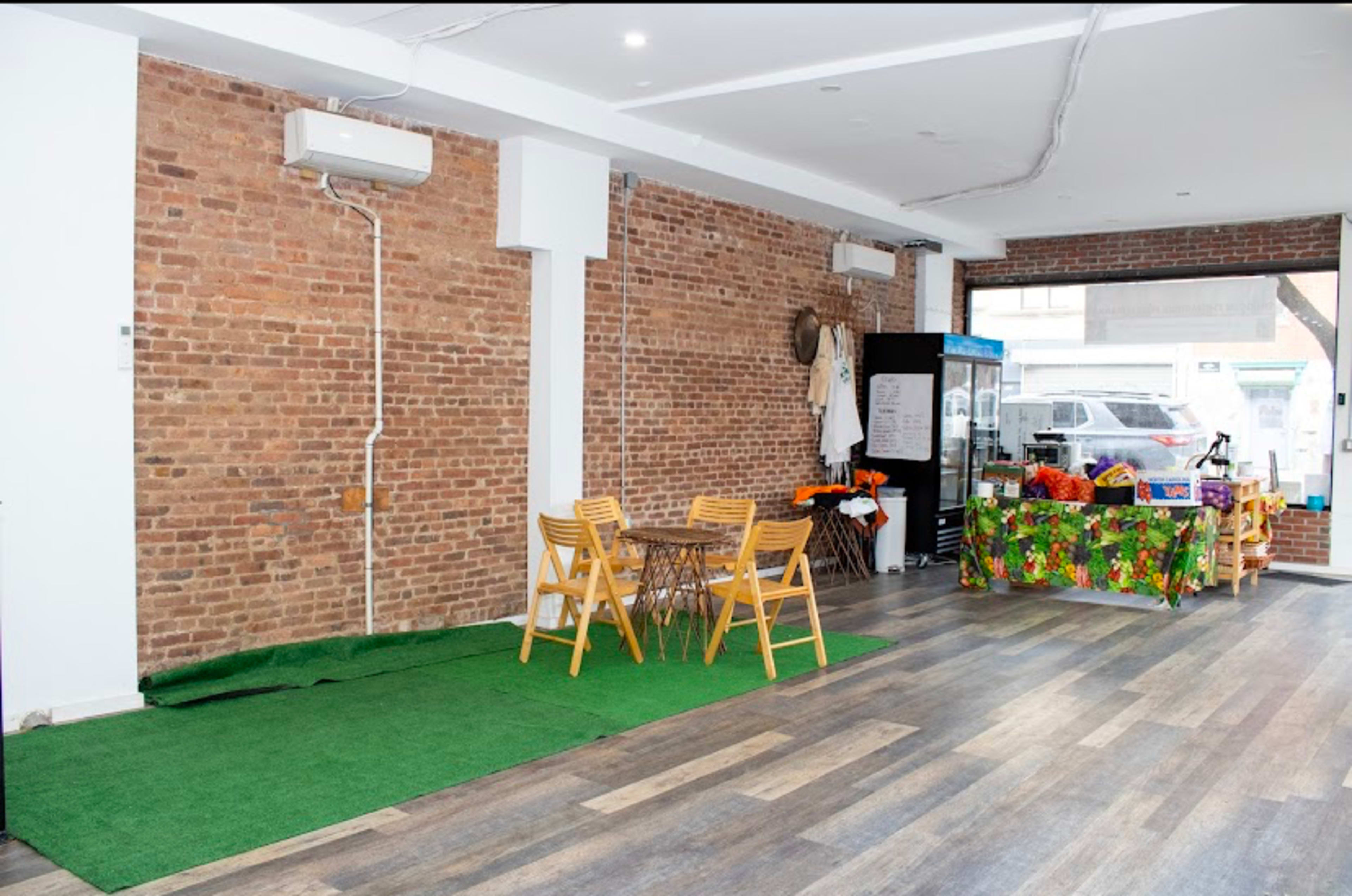 The image shows a spacious room with exposed brick walls, wooden flooring, a small area of artificial grass, a set of four wooden chairs, and a table covered in a colorful cloth.