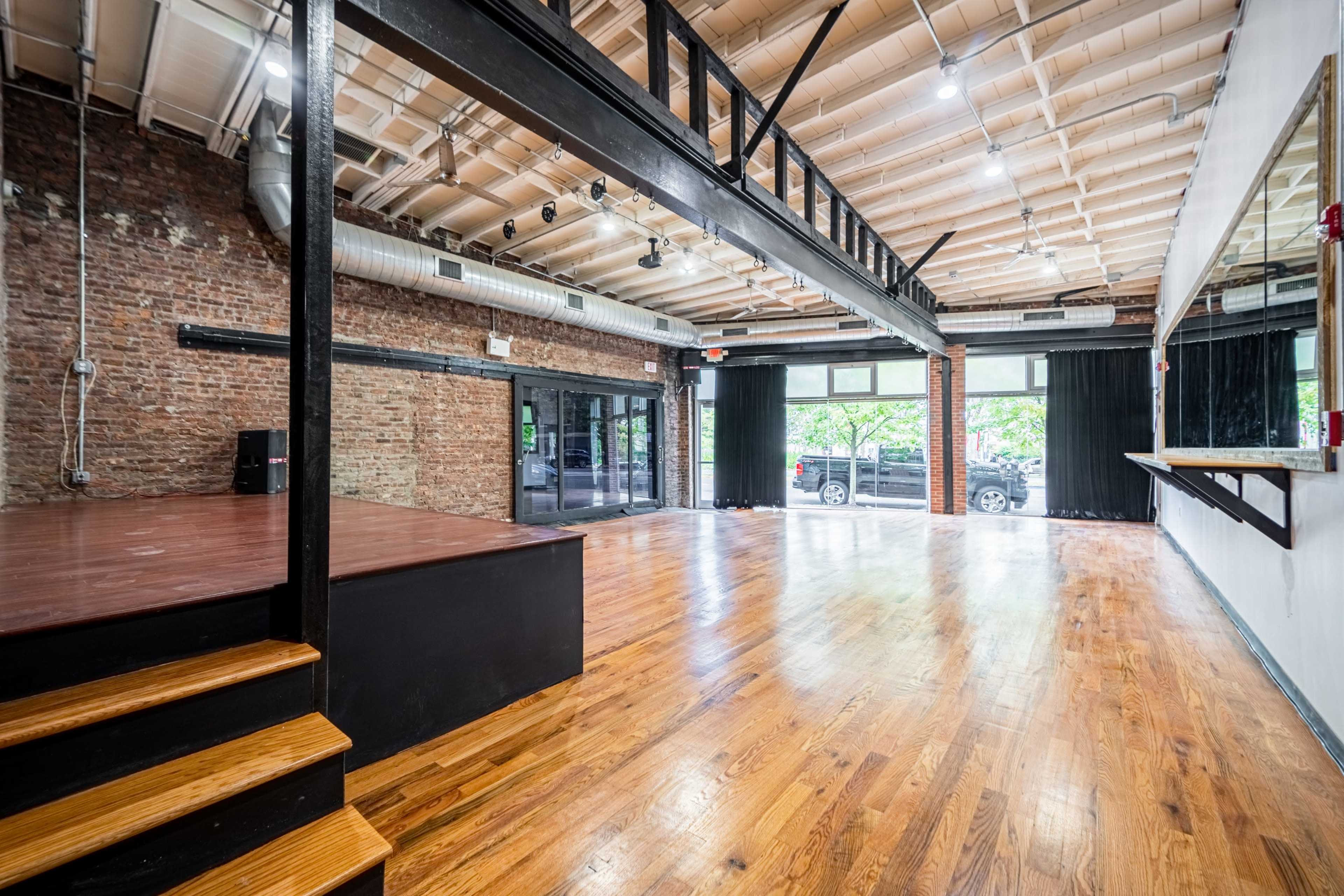 An empty interior space with wooden floors, exposed brick walls, large windows, and a raised platform on one side.