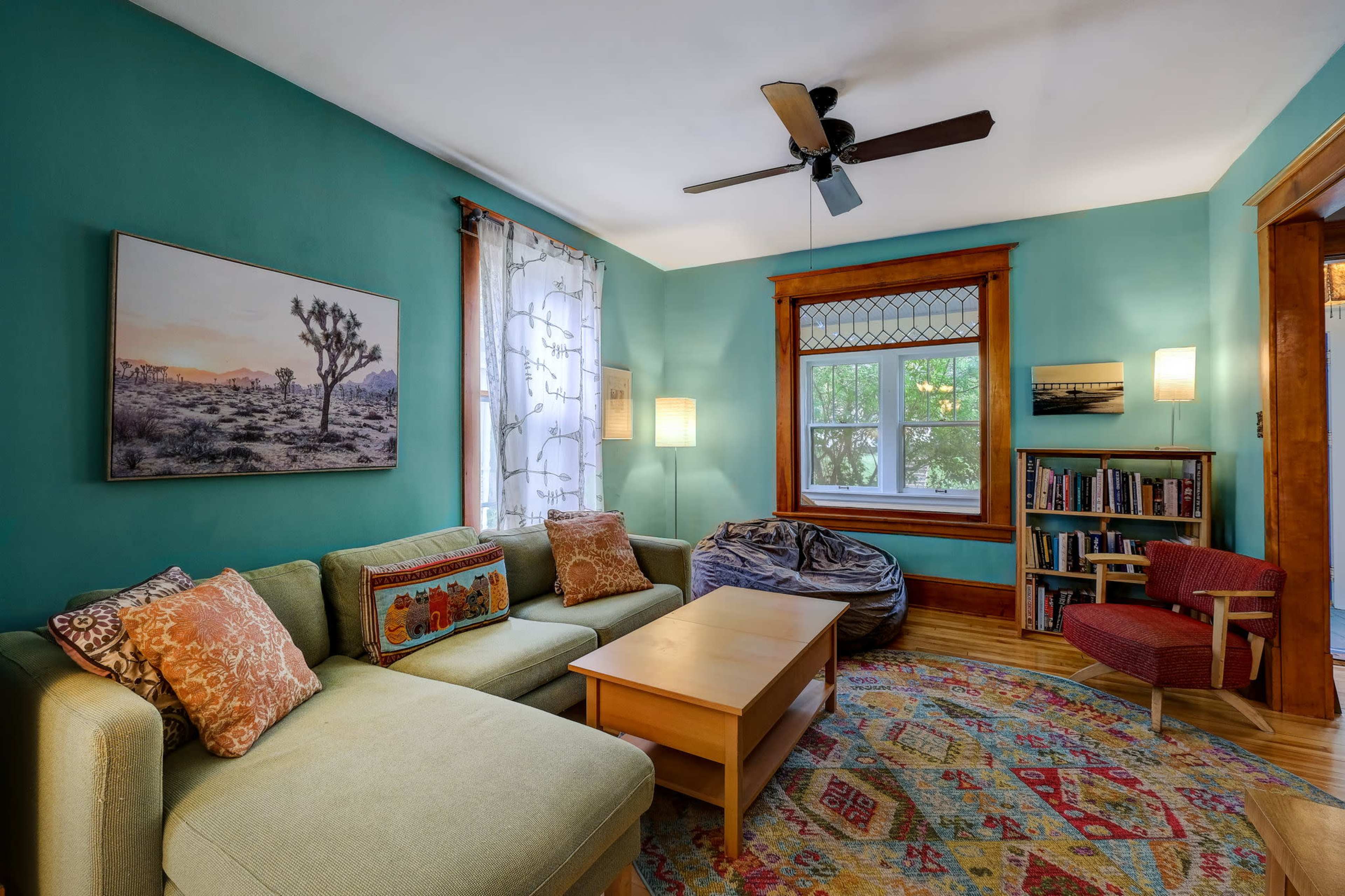 A cozy living room featuring a green wall, a sectional sofa with decorative pillows, a wooden coffee table, and a window with patterned curtains, accompanied by bookshelves and a bean bag chair.