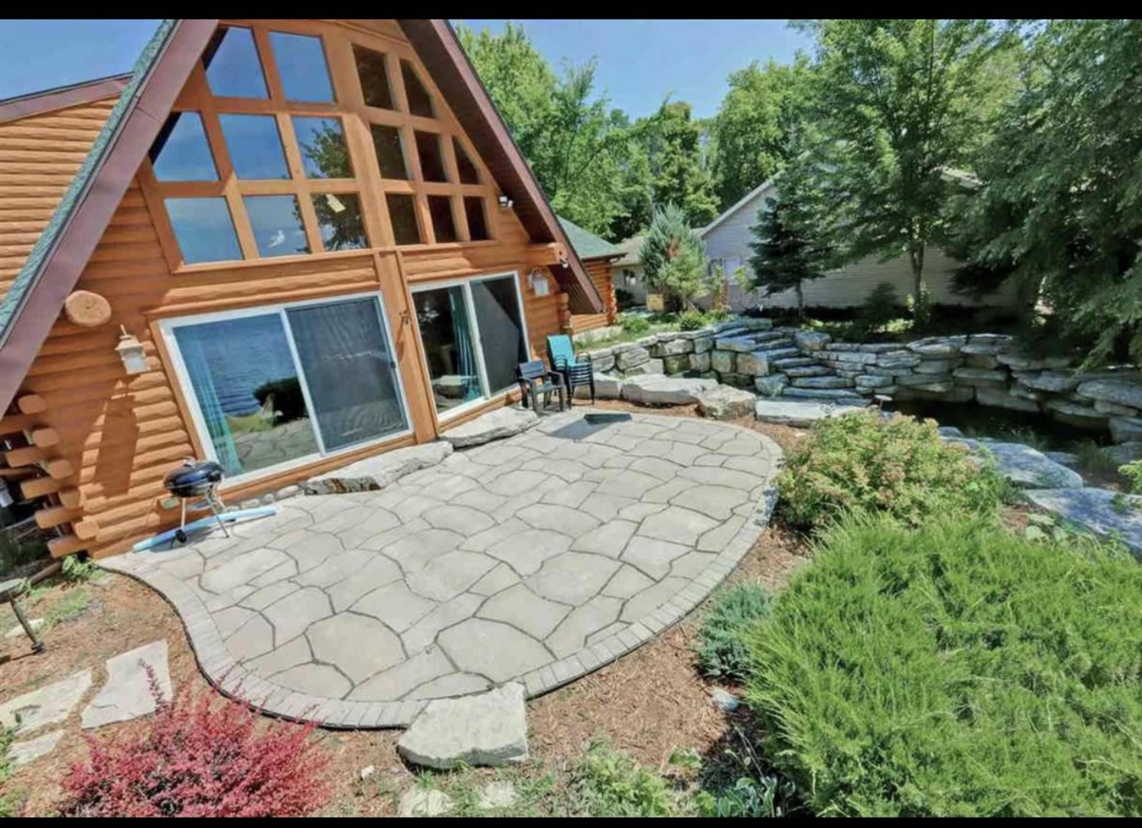 A stone patio with a grill is situated outside a house featuring large windows and an A-frame design, adjacent to a landscaped area with rocks and greenery.
