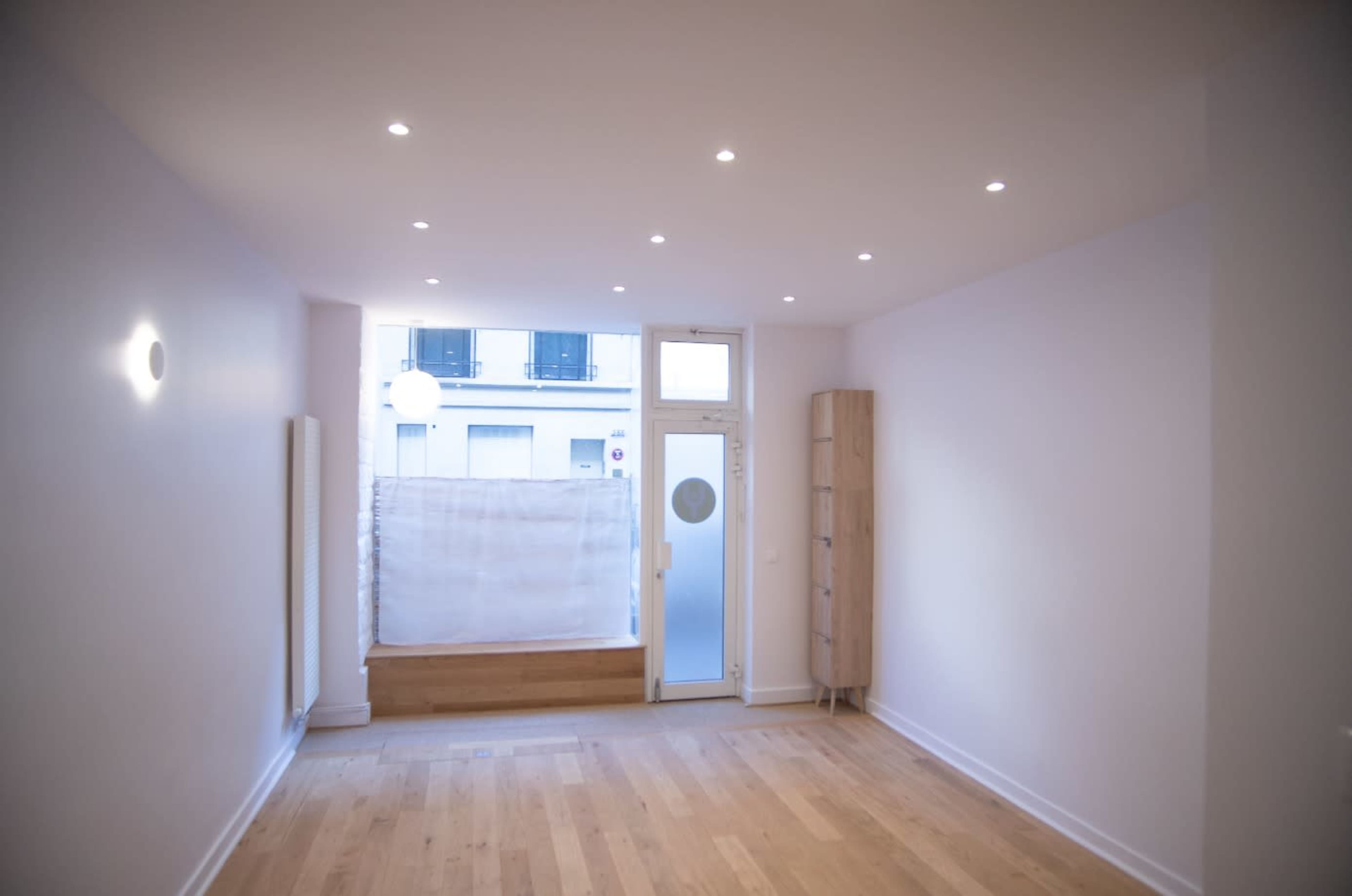An empty room with wooden flooring, a large window with a partially drawn curtain, and a simple wooden cabinet against one wall.