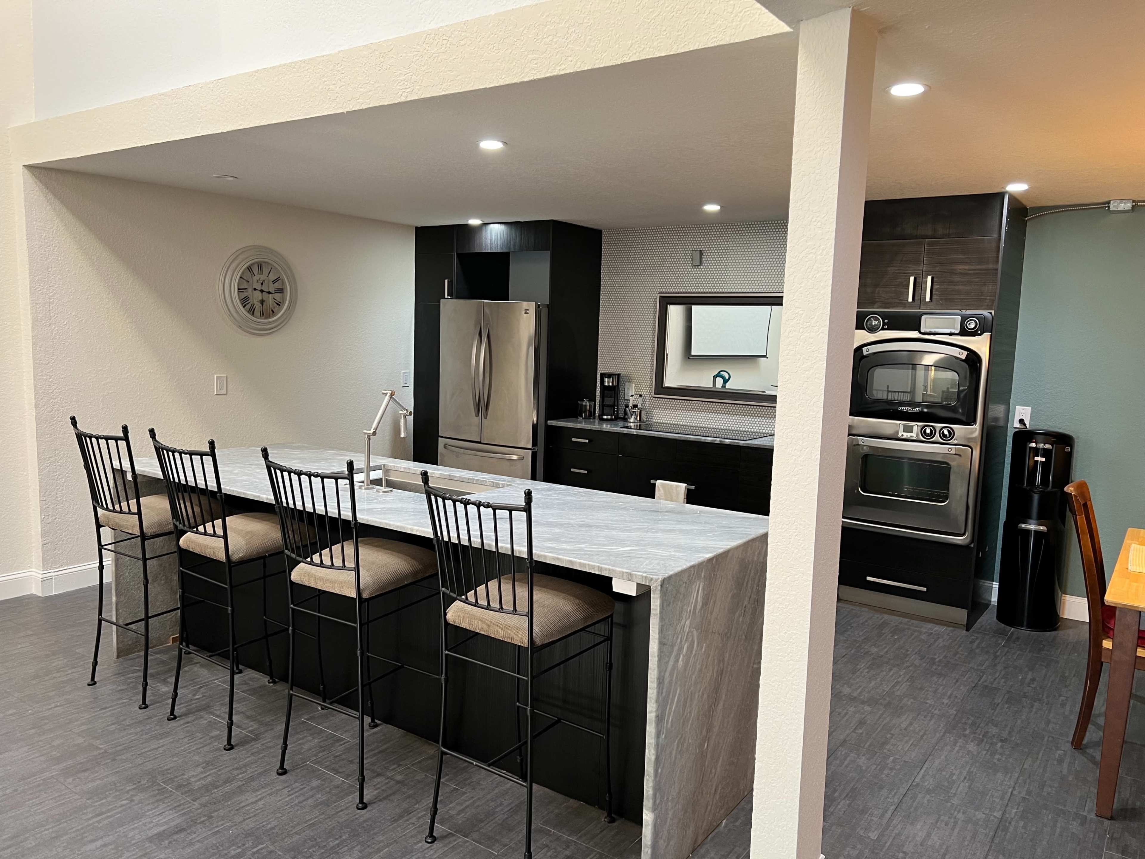 A modern kitchen with a long, gray stone island and black cabinetry, featuring a stainless steel refrigerator and an oven.