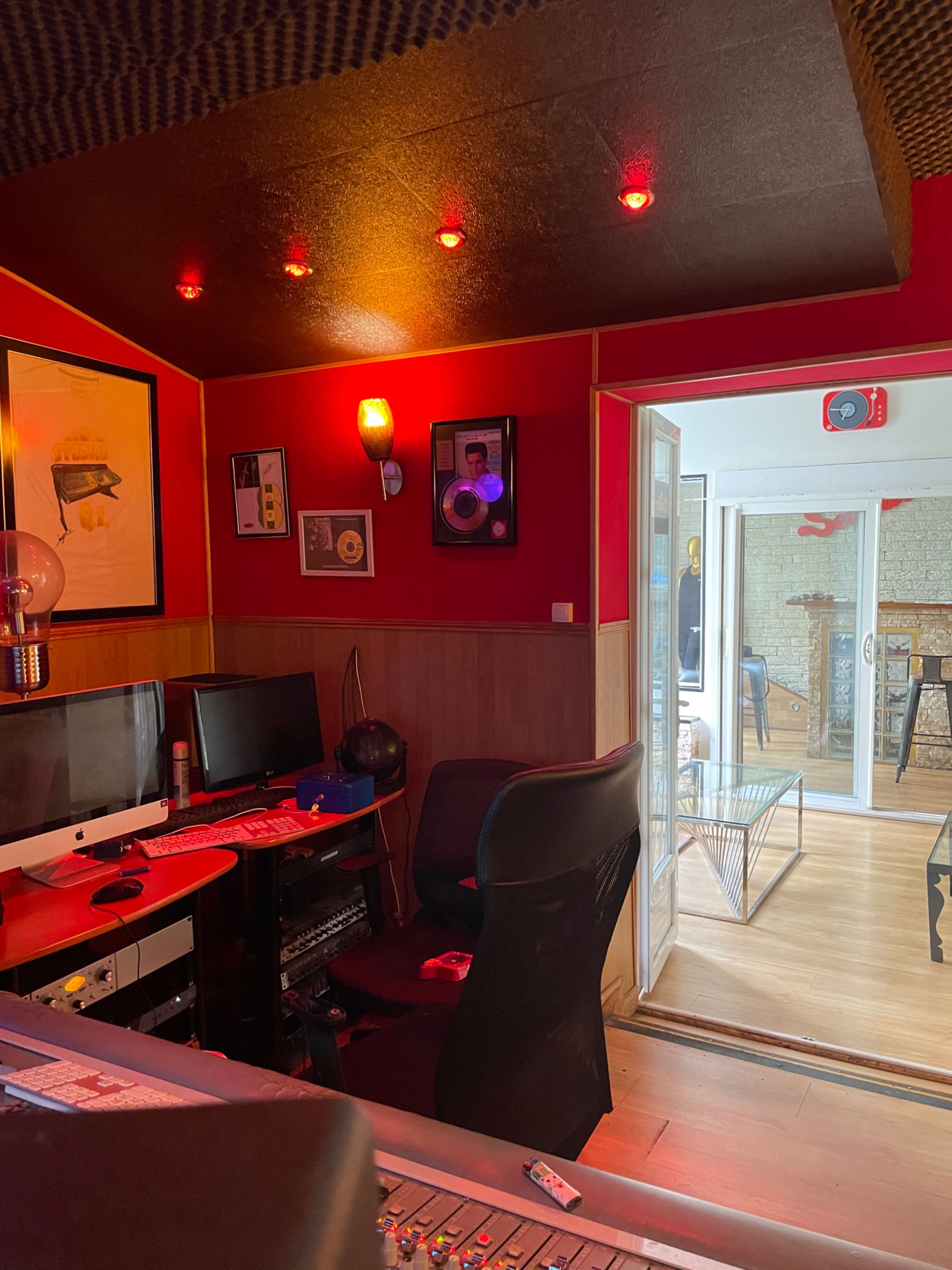 A dimly lit recording studio with red walls, a mixing console in the foreground, and a glass door leading to a bright, multicolored space.