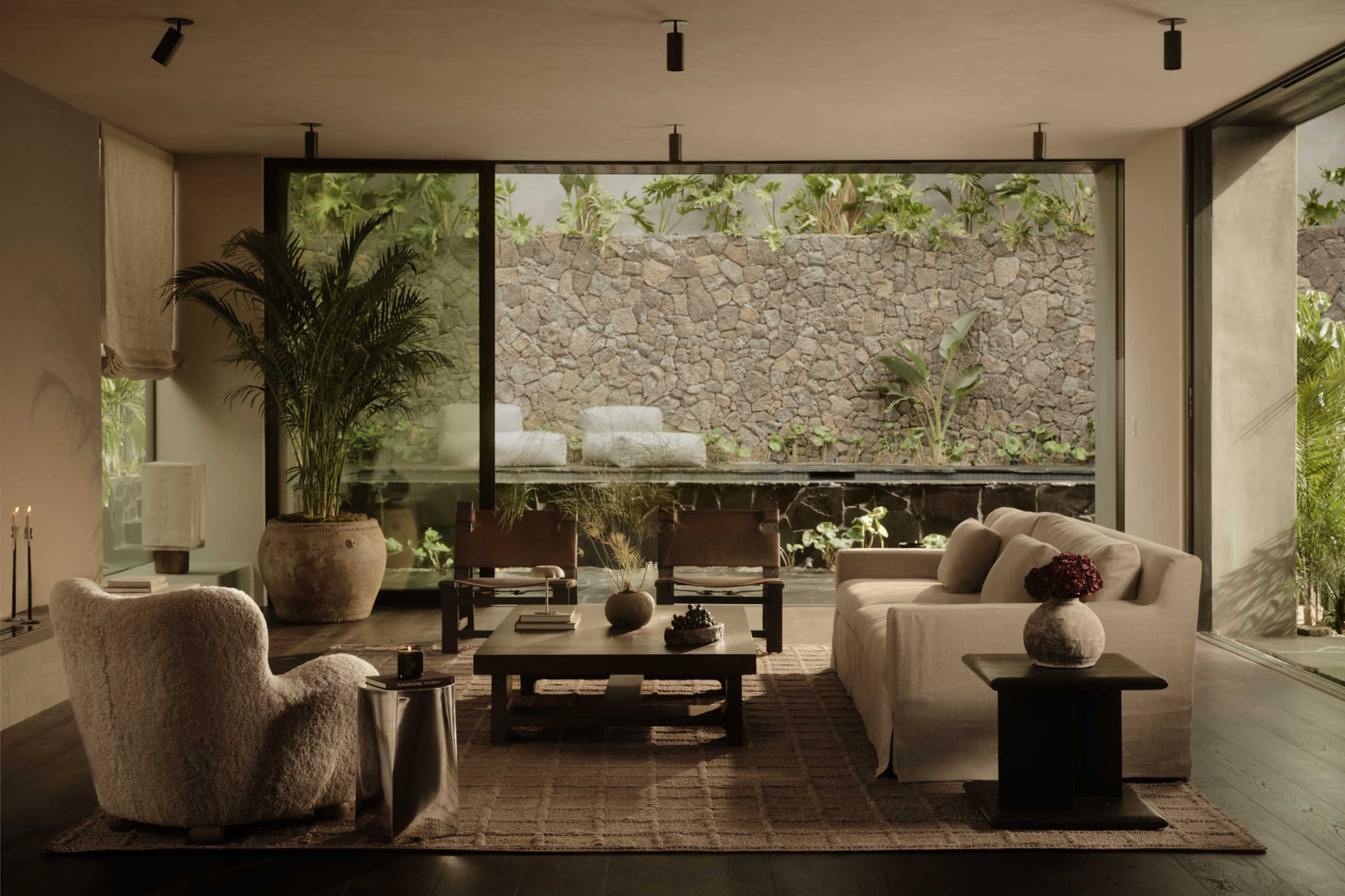 The image depicts a modern living room with large glass windows overlooking a stone wall garden, featuring minimalist furniture including a white sofa, armchairs, and a coffee table.