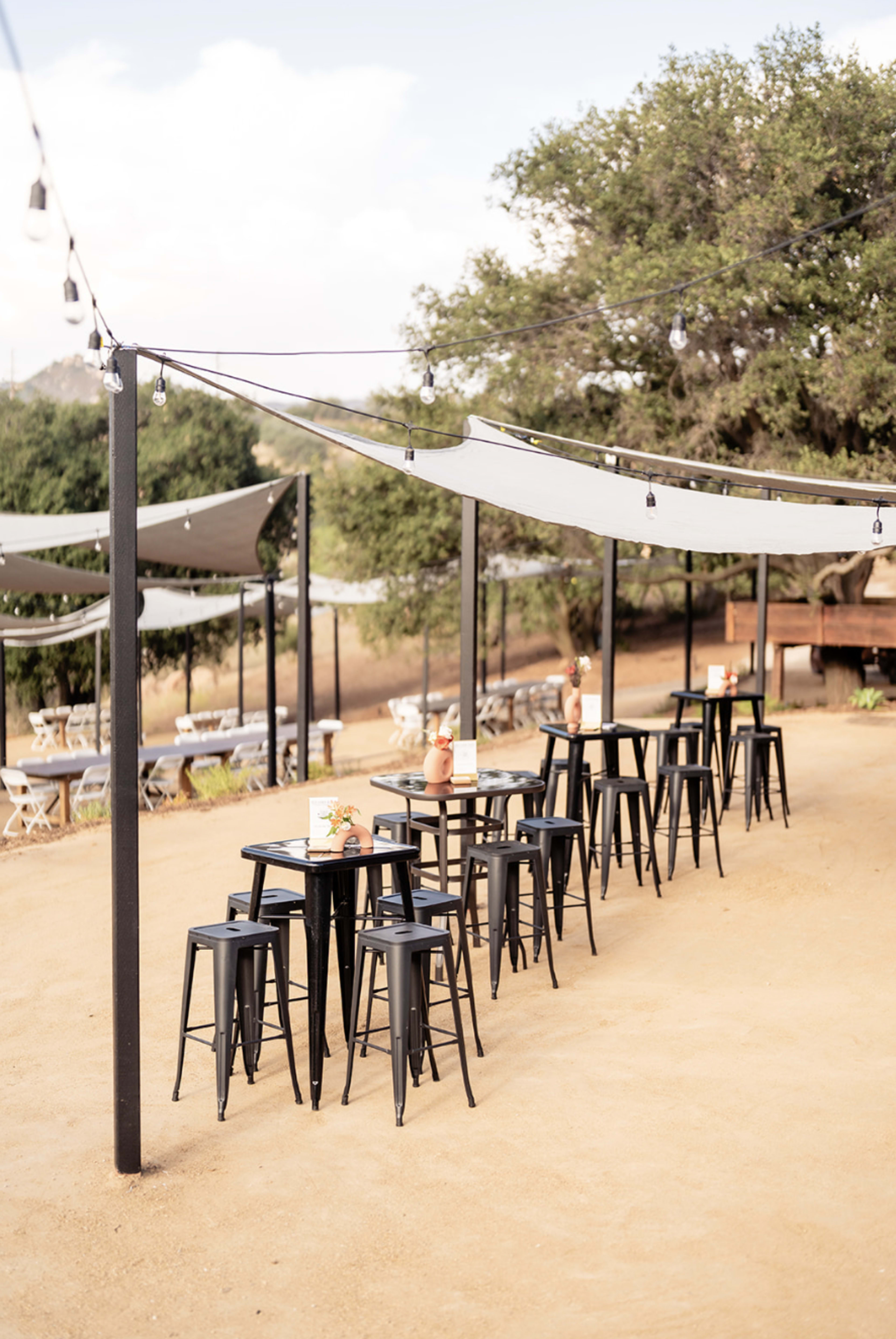 An outdoor dining area features rows of tall black stools and tables under shaded canopies, surrounded by trees.