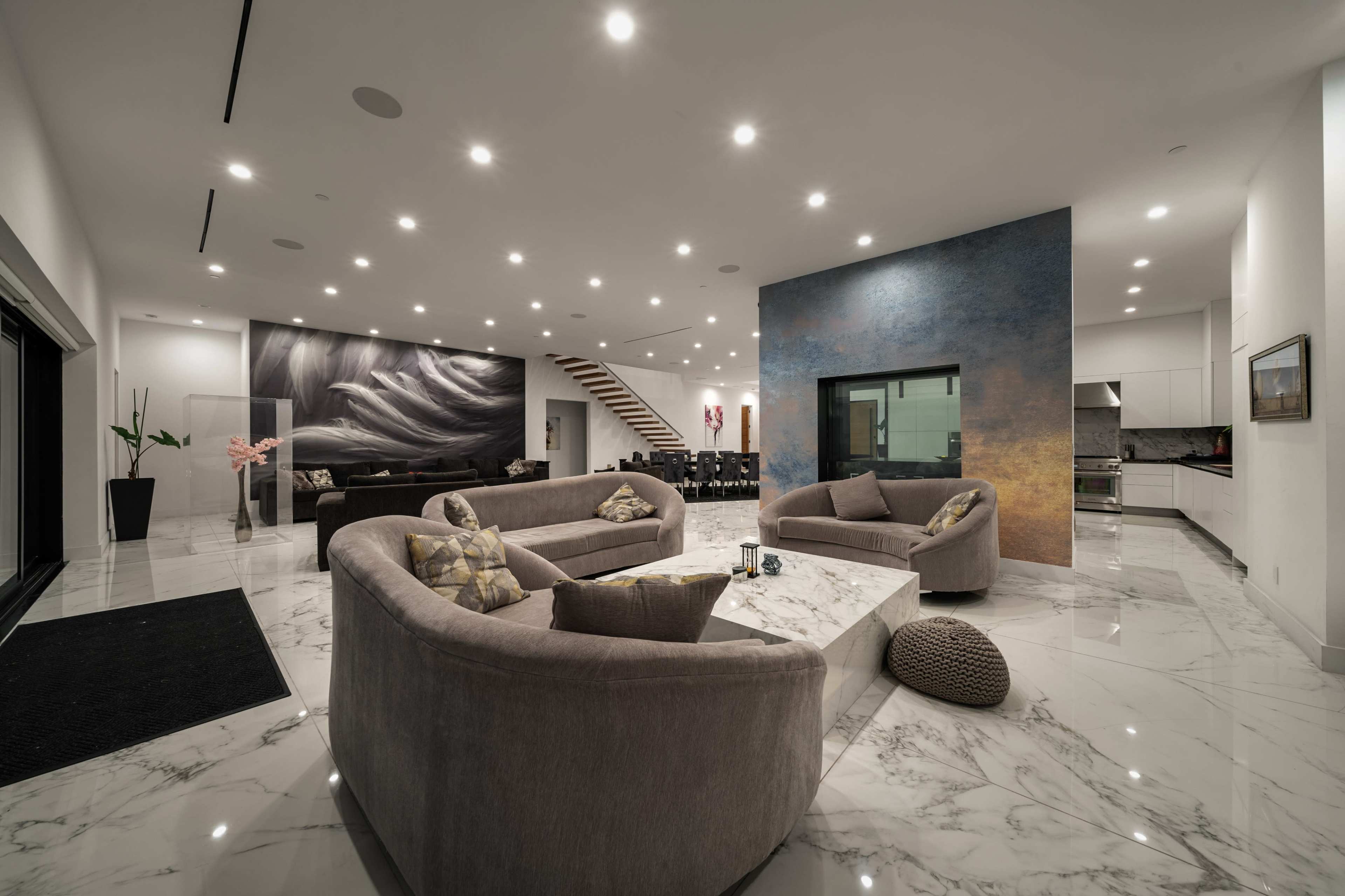 A modern living area with marble flooring, two large gray sofas arranged around a coffee table, and a staircase in the background leading to a dining area.