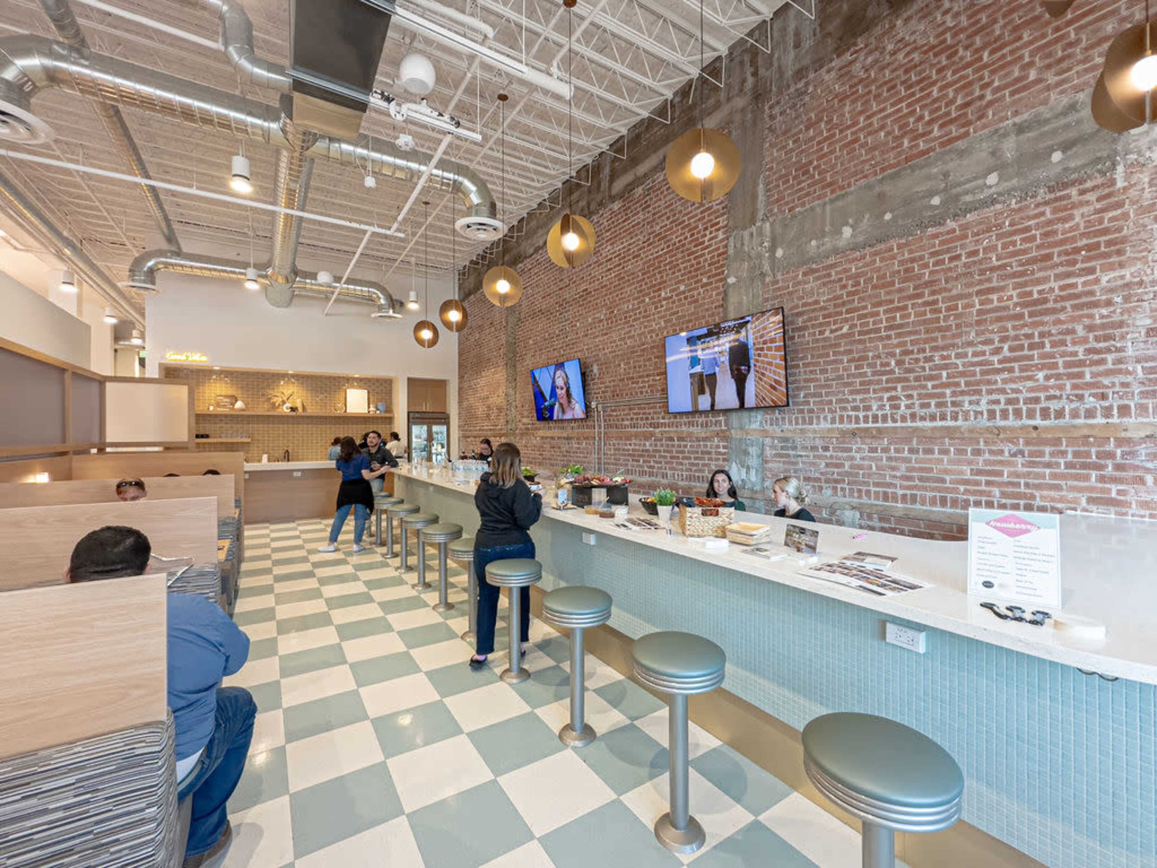 The image shows a modern cafe interior featuring a bar with high stools, exposed brick walls, and large screens displaying videos.