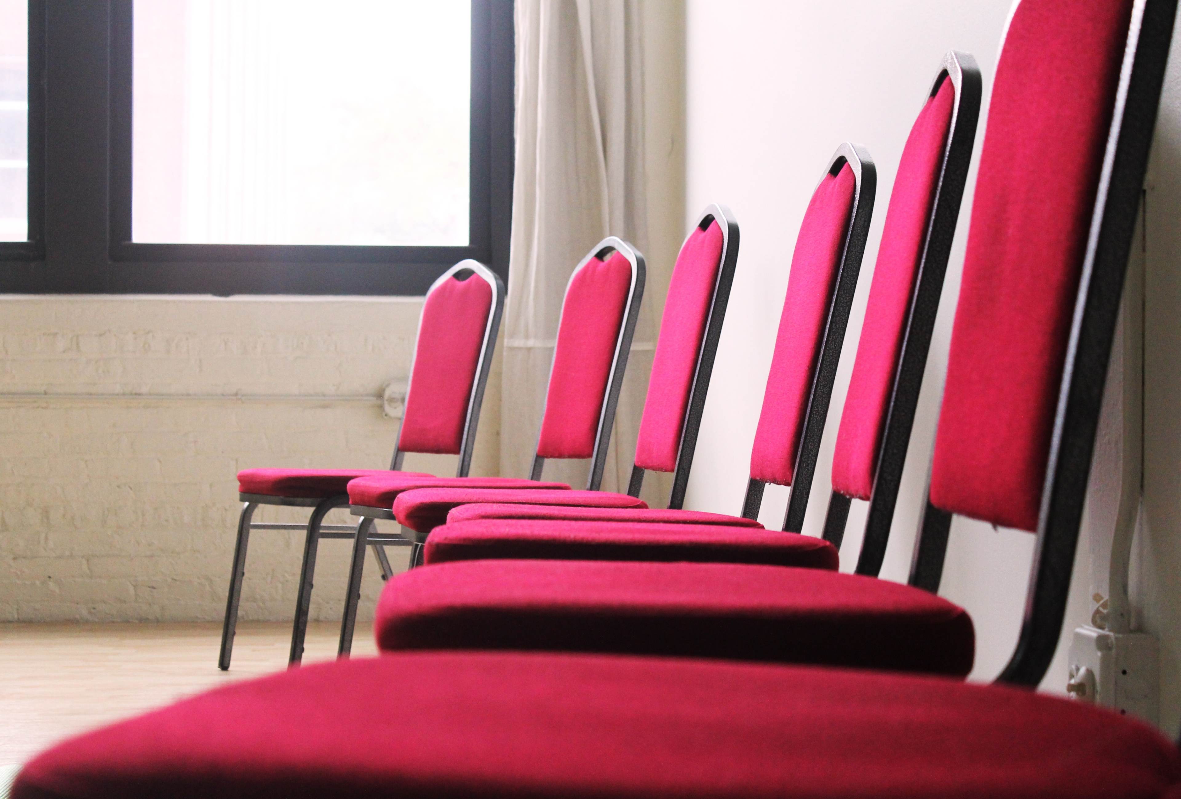 A row of five red chairs with metal frames is positioned against a wall near a window.
