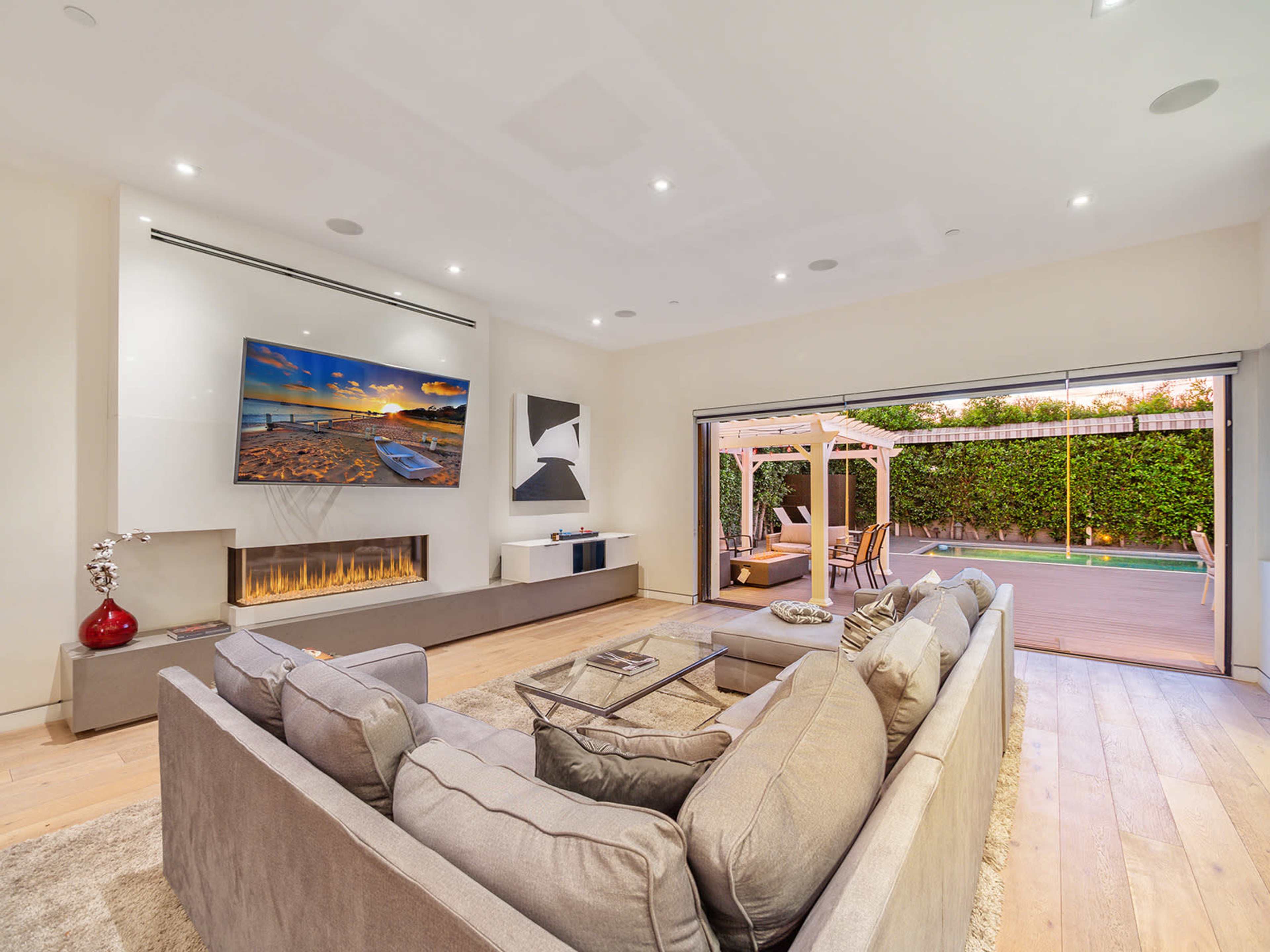 A modern living room features a large wall-mounted TV above a sleek fireplace, with a sectional sofa facing a wide sliding door that opens to an outdoor patio.