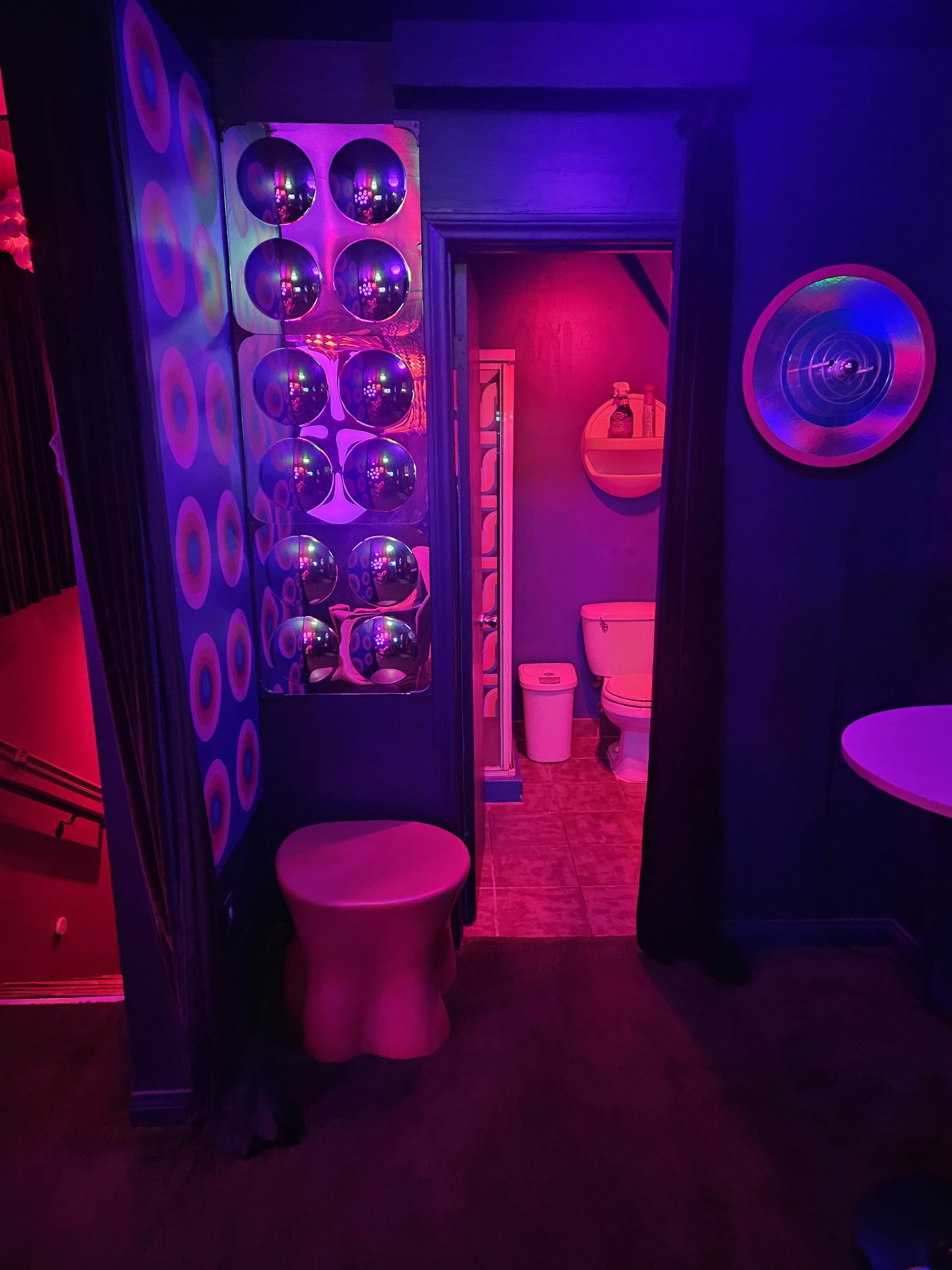 The image shows a dimly lit room with purple and red lighting, featuring a small bathroom area with a toilet and a wall covered in reflective circular decorations.