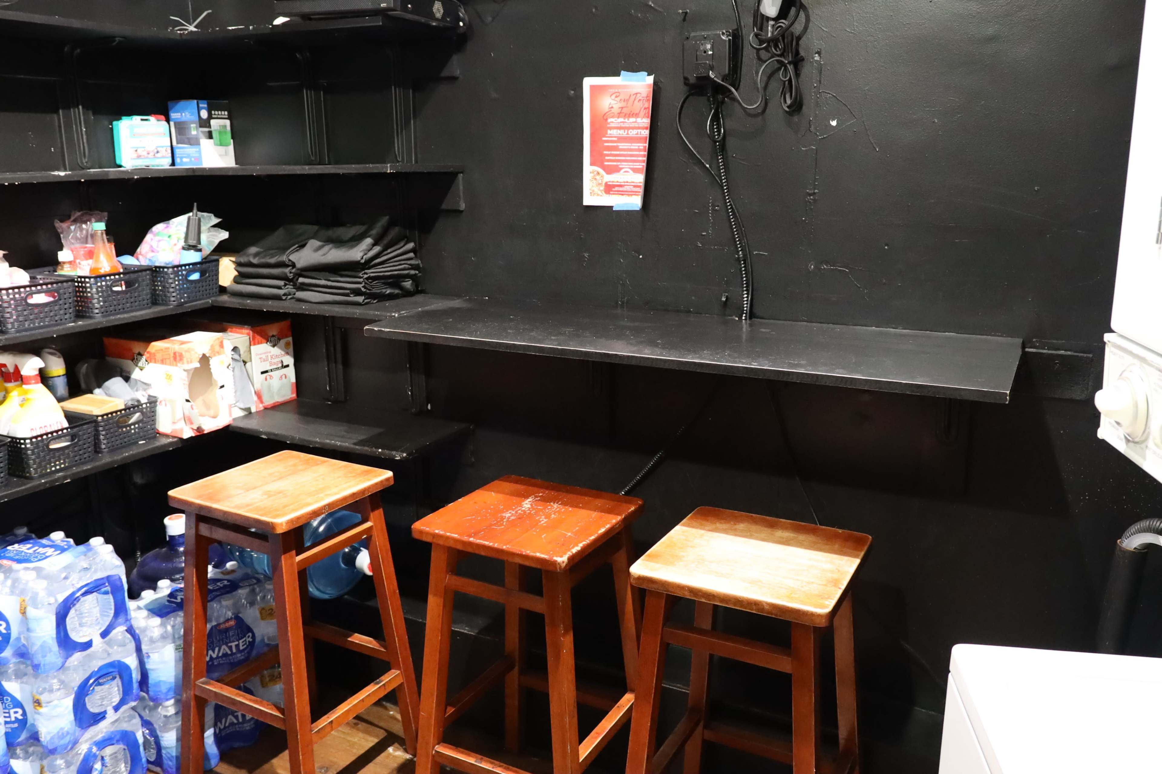 A small storage room with black walls, featuring three wooden stools in front of a black shelf and various items on adjacent shelves.