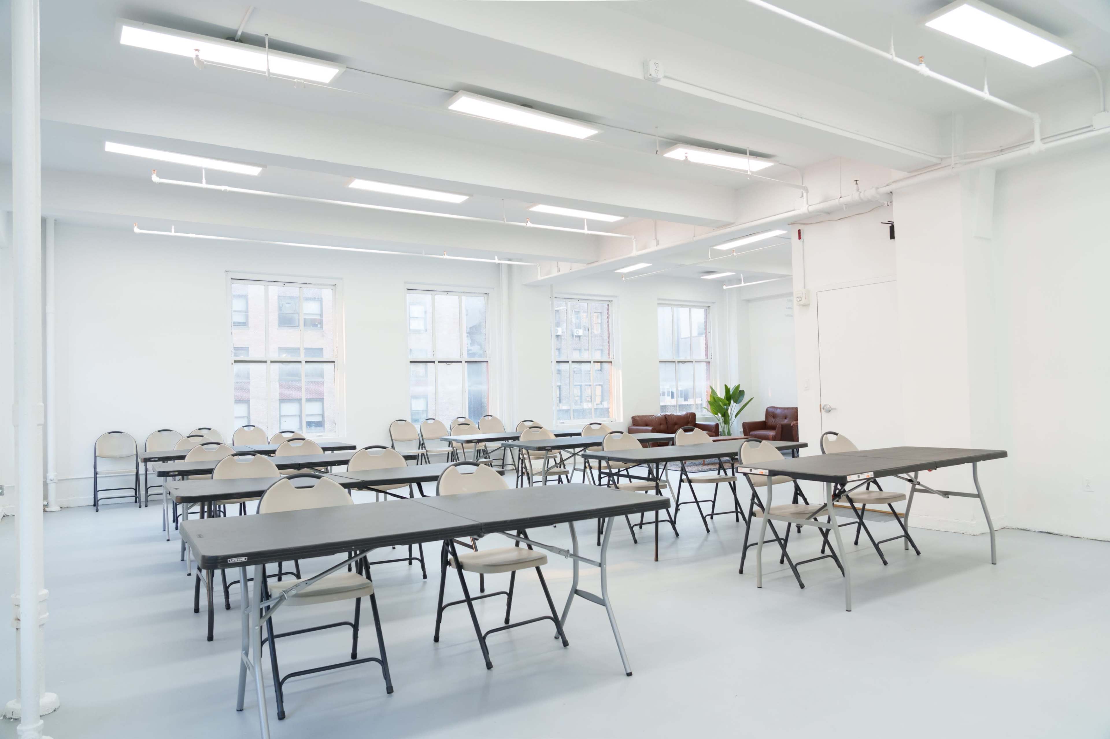 The image shows a spacious, well-lit room arranged with multiple tables and chairs, with two armchairs and a plant in one corner.
