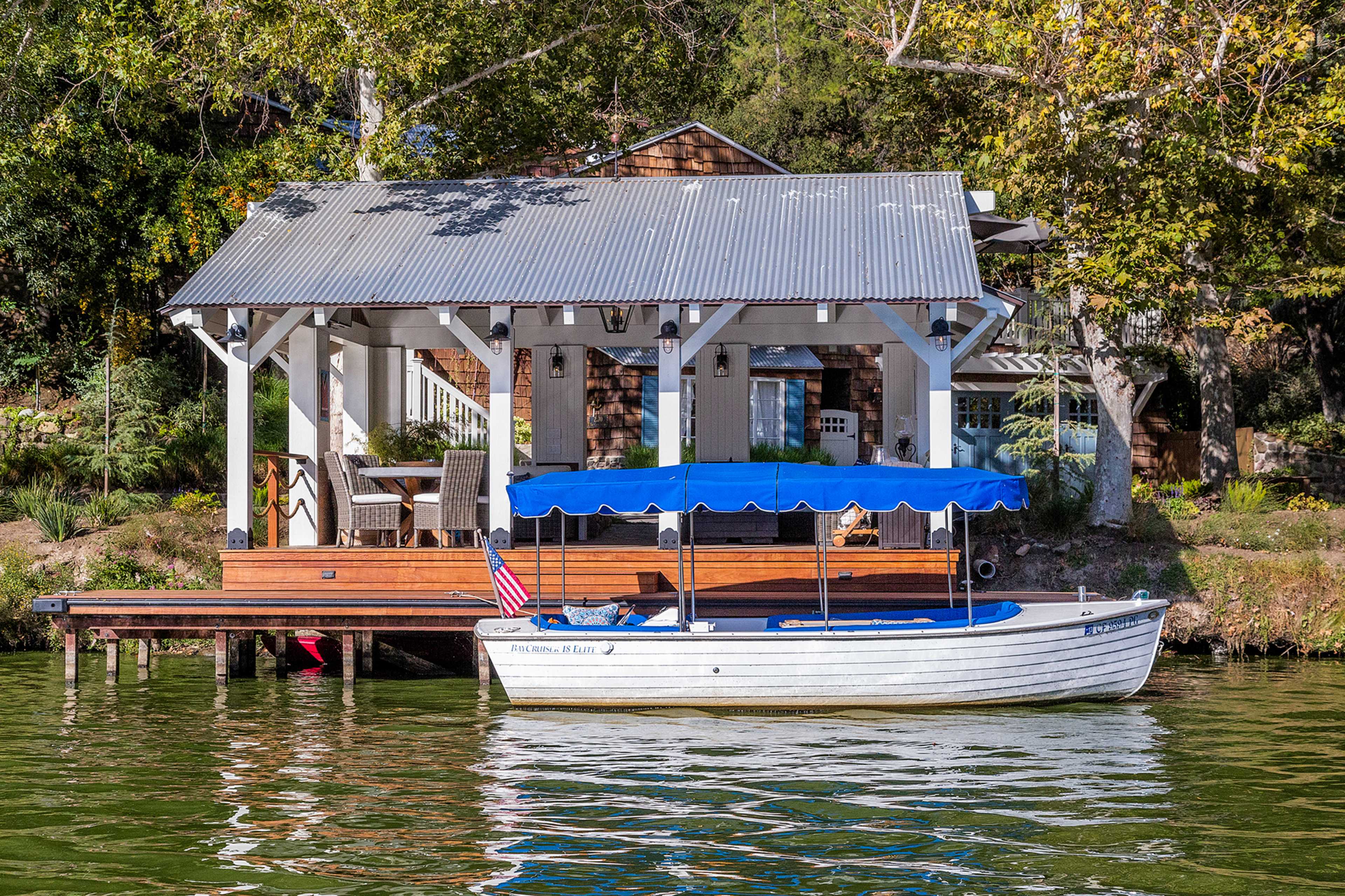 A white boat with a blue canopy is docked beside a riverside pavilion featuring a metal roof and wooden deck.