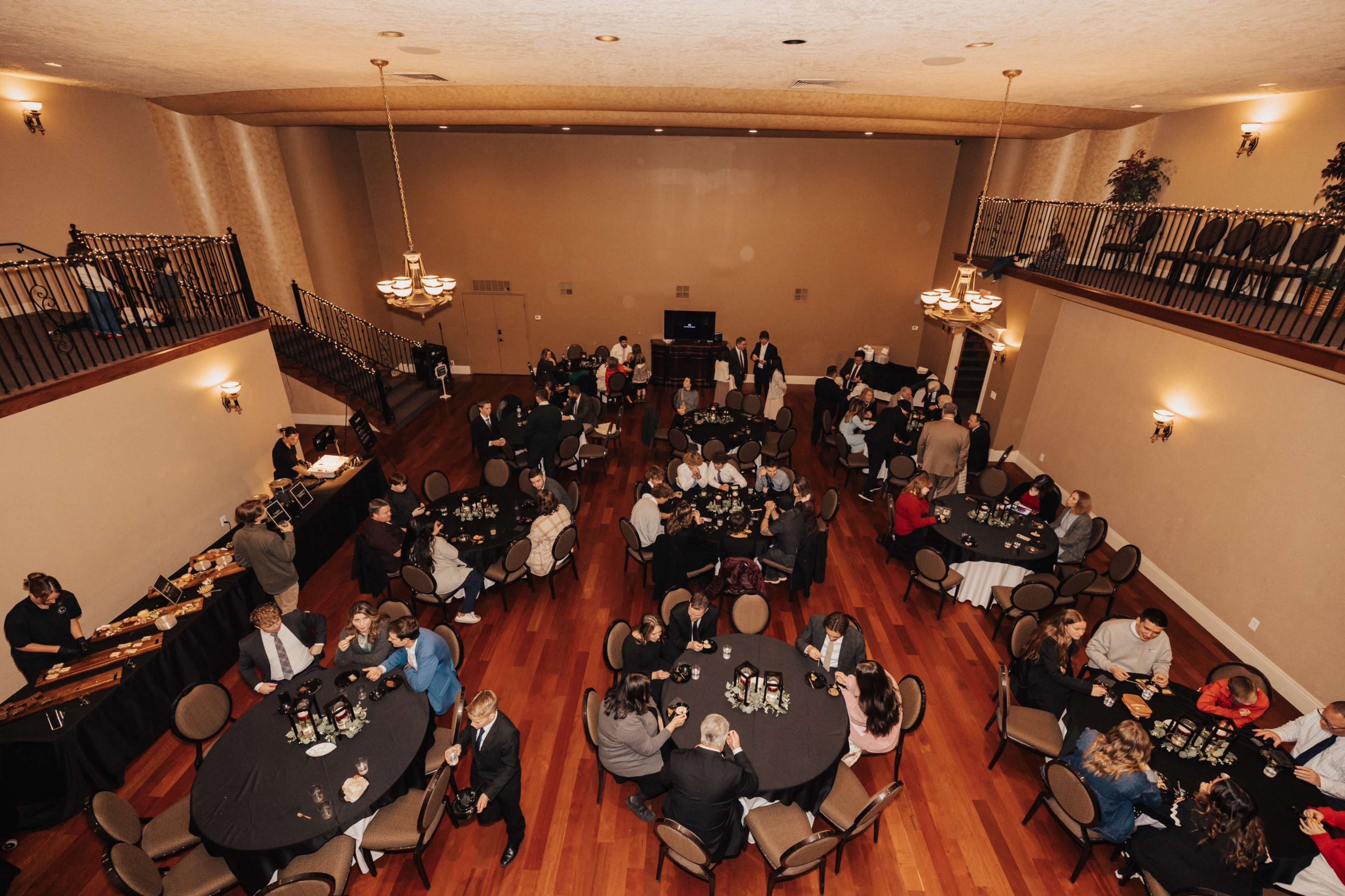 A large banquet hall features several round tables occupied by guests, with a buffet setup and an elevated stage in the background.