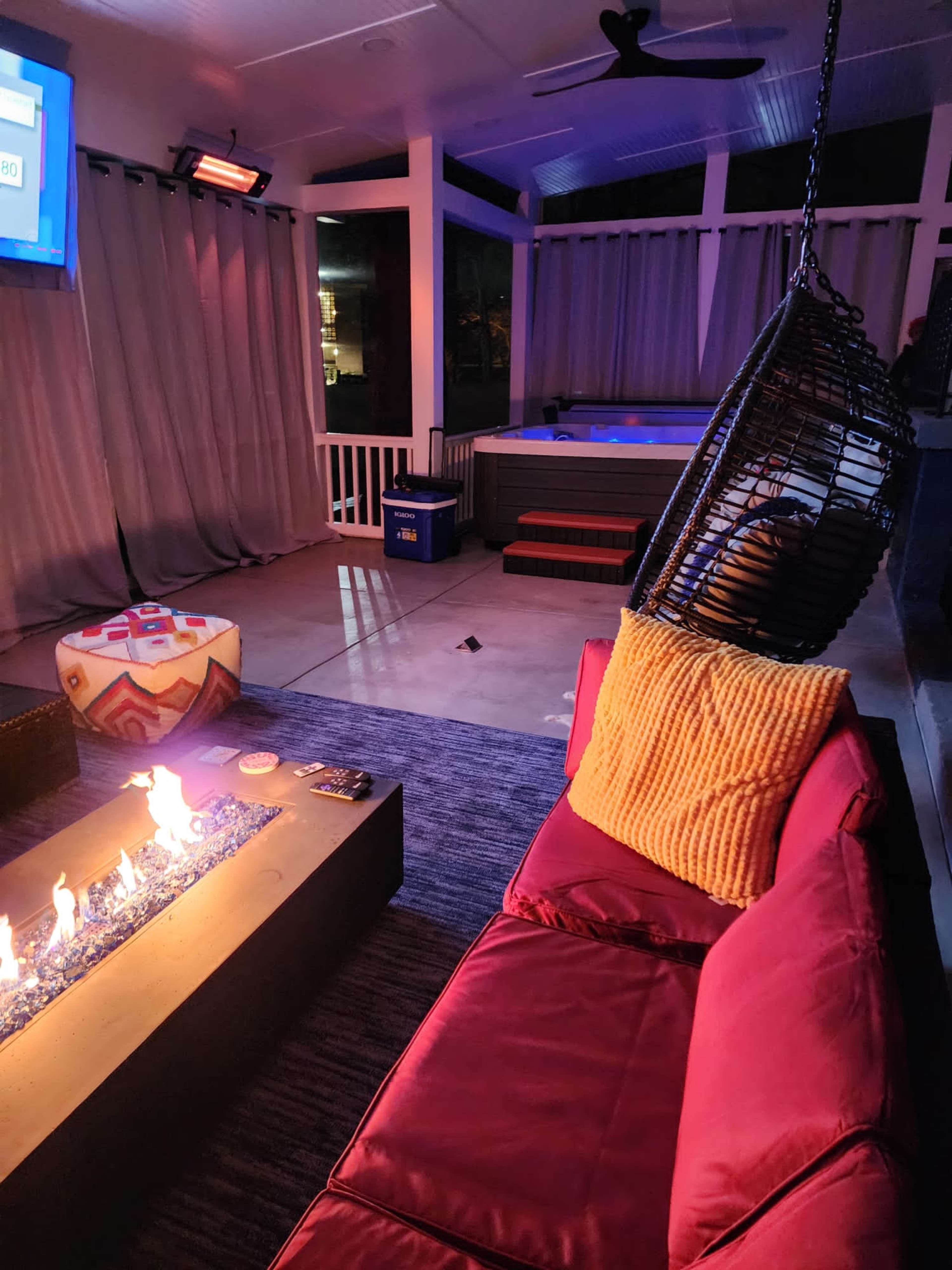 A covered outdoor lounge area with a fire pit, a hanging chair, and a hot tub in the background.