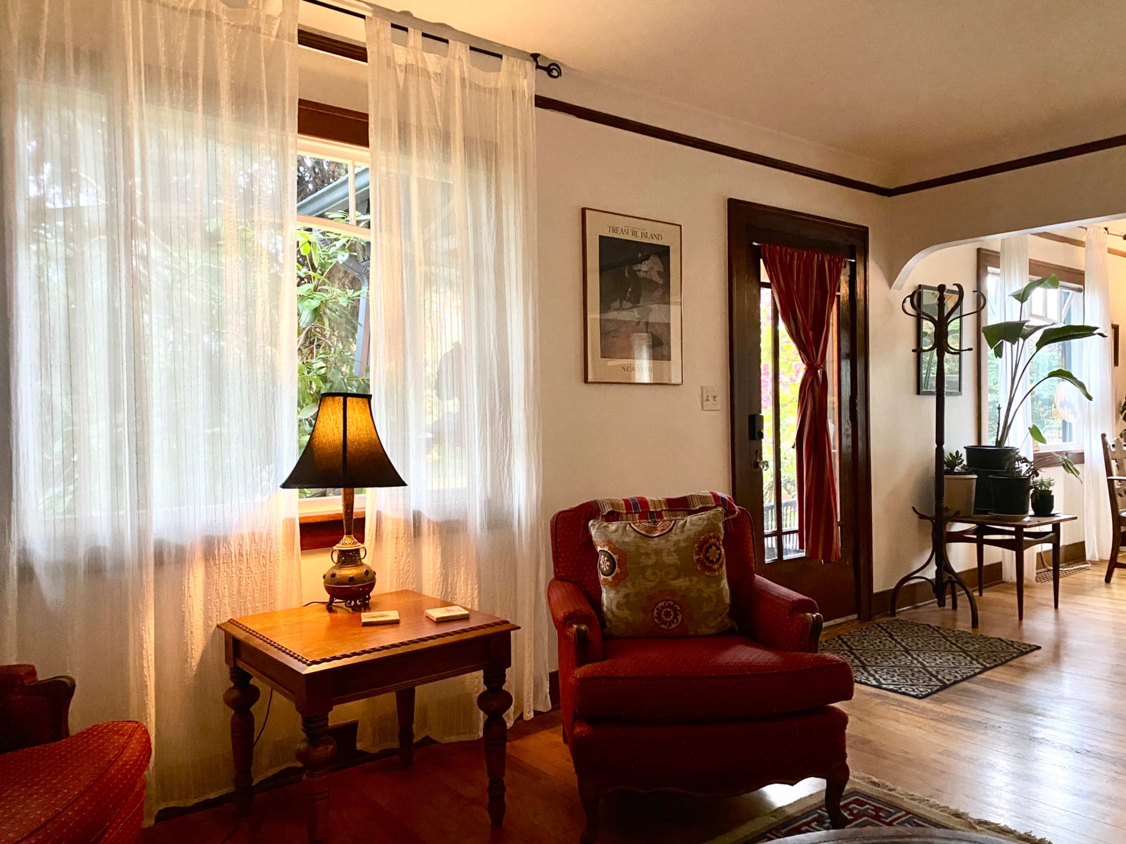 A cozy living room with a lamp on a side table, a comfortable chair, sheer curtains, and a view of an entrance with a plant nearby.