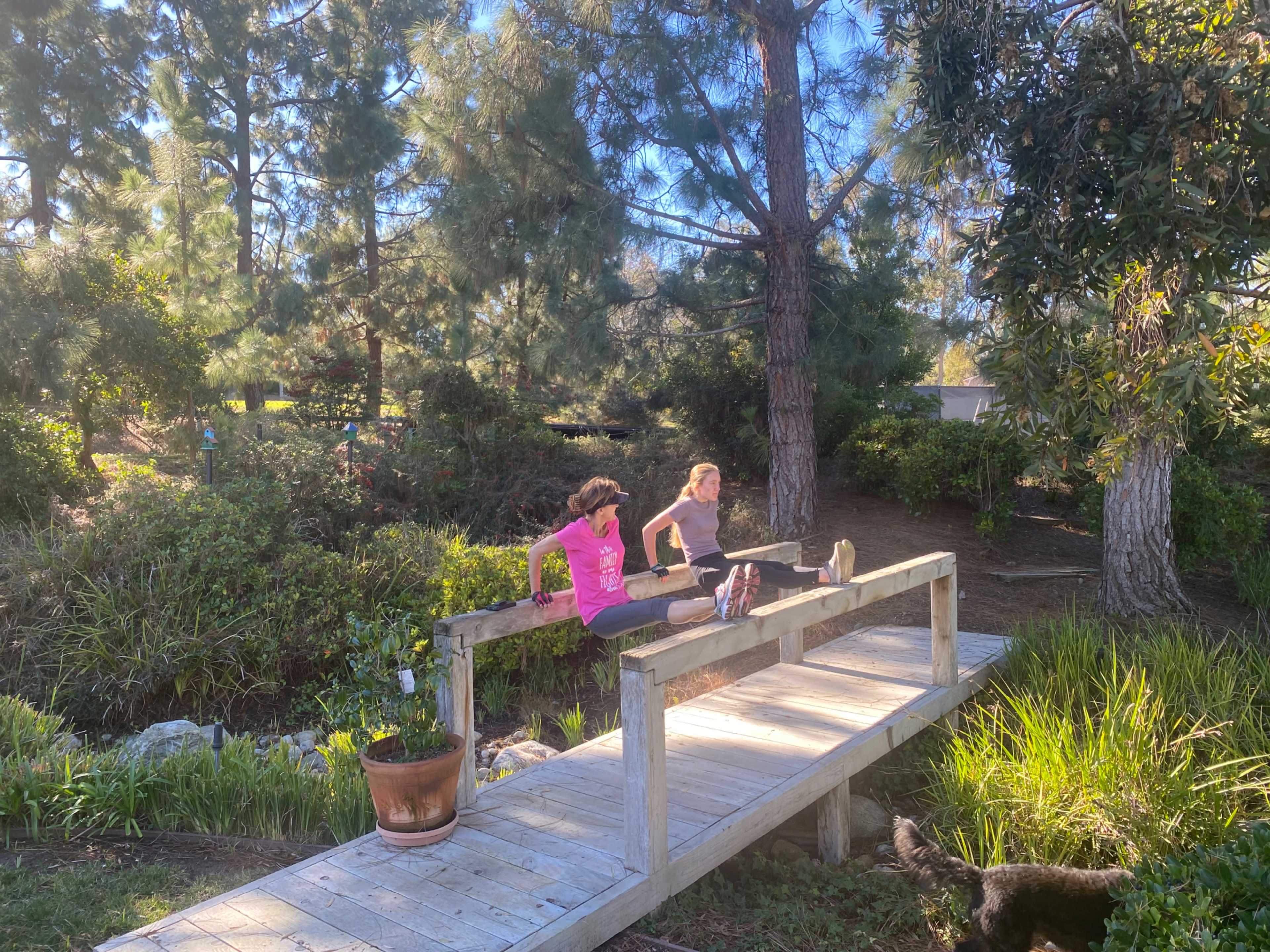 Two women exercise on a wooden bridge in a park surrounded by trees and greenery.