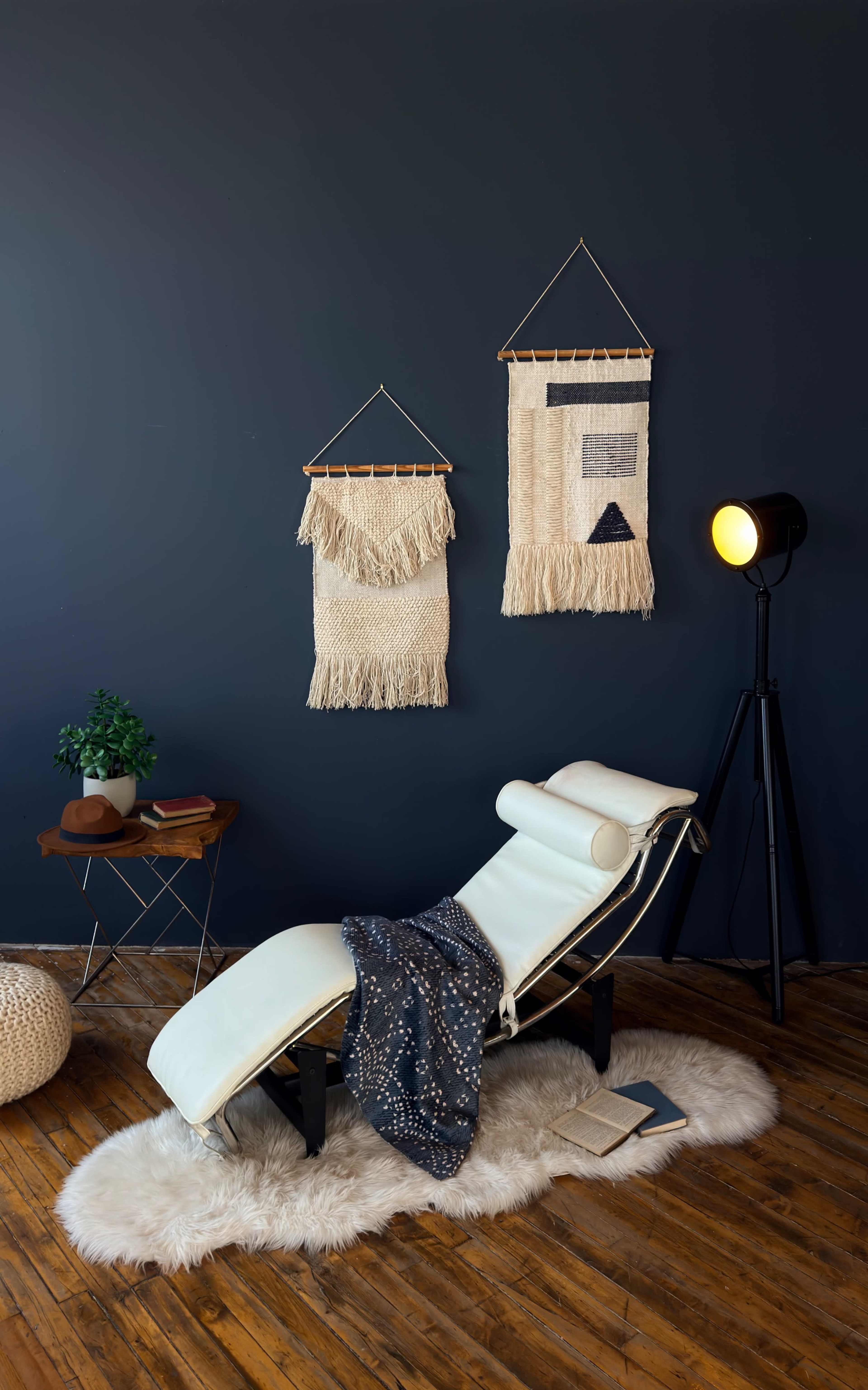 The image shows a modern lounge chair with a cozy blanket draped over it, set against a dark wall decorated with woven wall hangings, accompanied by a floor lamp and a small side table.