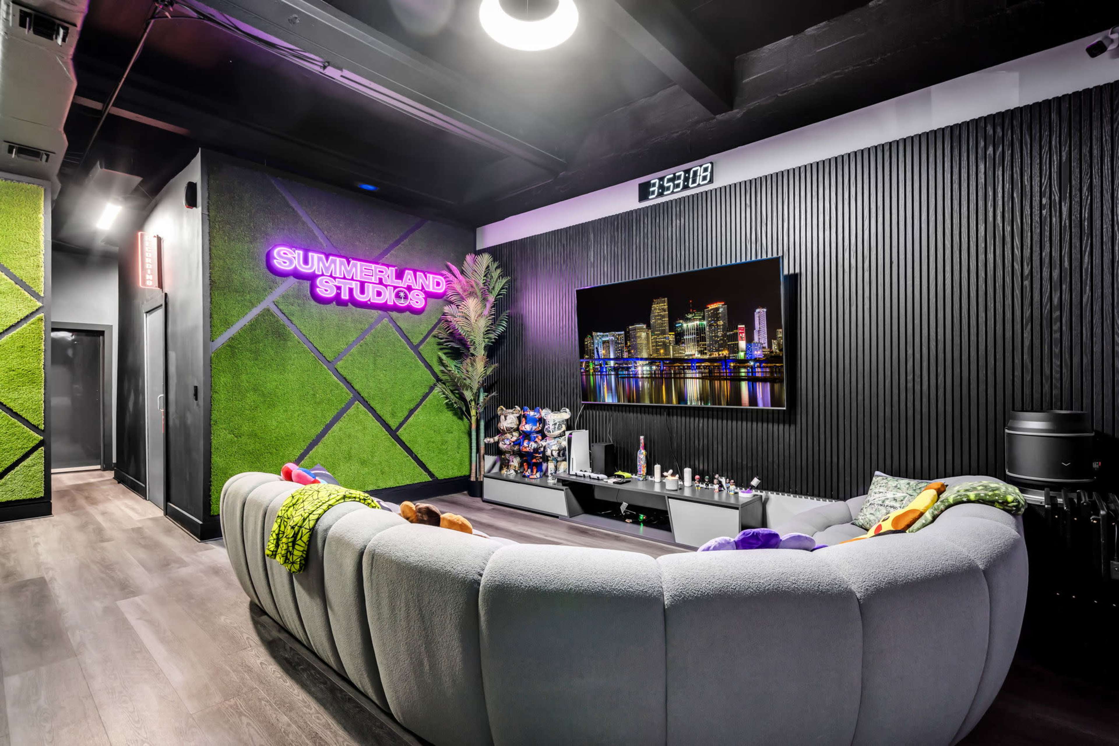 The image features a modern studio lounge with a curved gray sofa, a large television mounted on a wall, and vibrant neon signage that reads "SUMMERLAND STUDIOS."