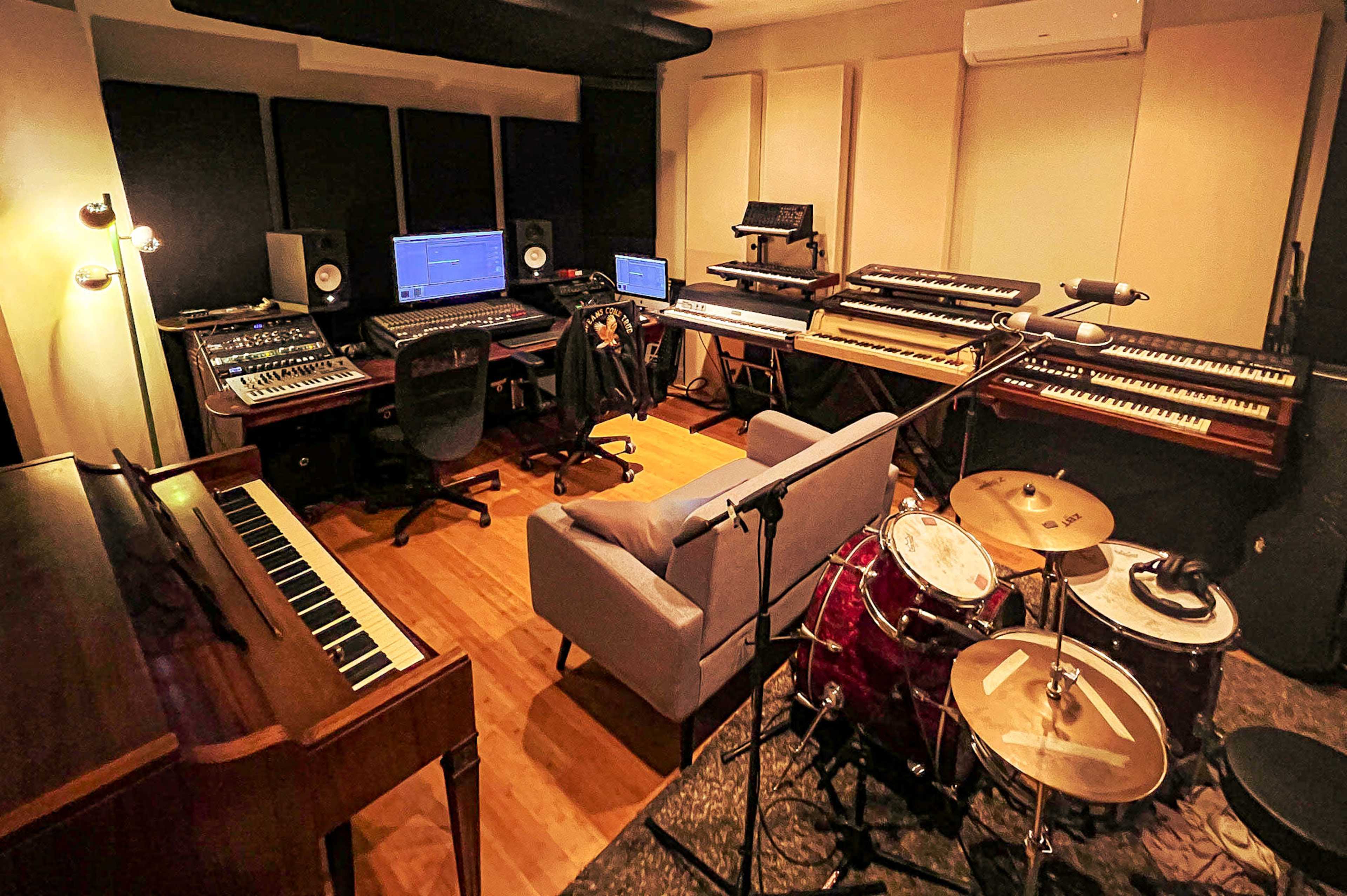 A music studio featuring a mixing console, multiple keyboards, a piano, a drum set, and seating arrangements.