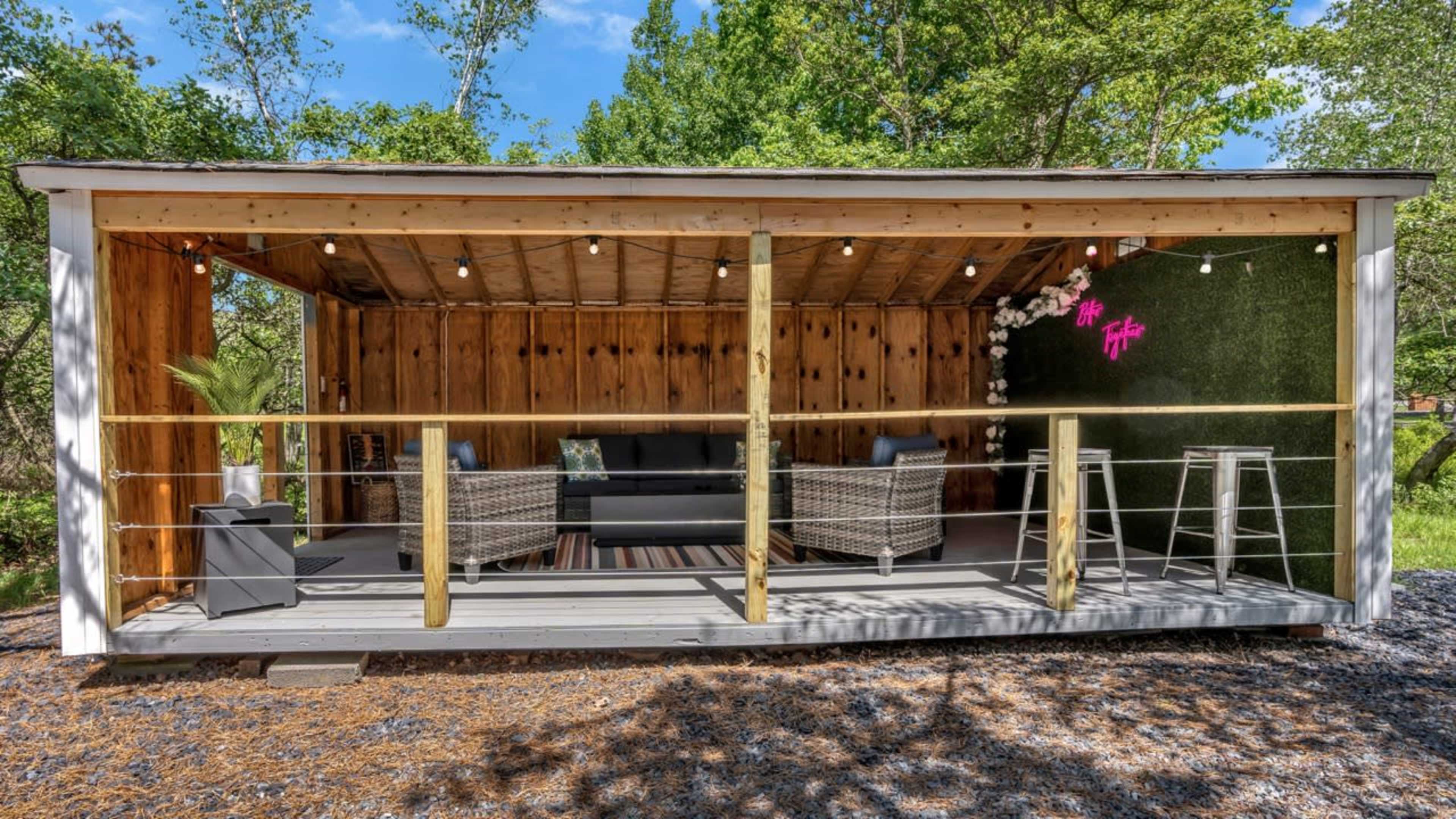 The image shows a modern outdoor lounge area with two wicker chairs, a sofa, and decorative plants under string lights, set within a wooden structure.
