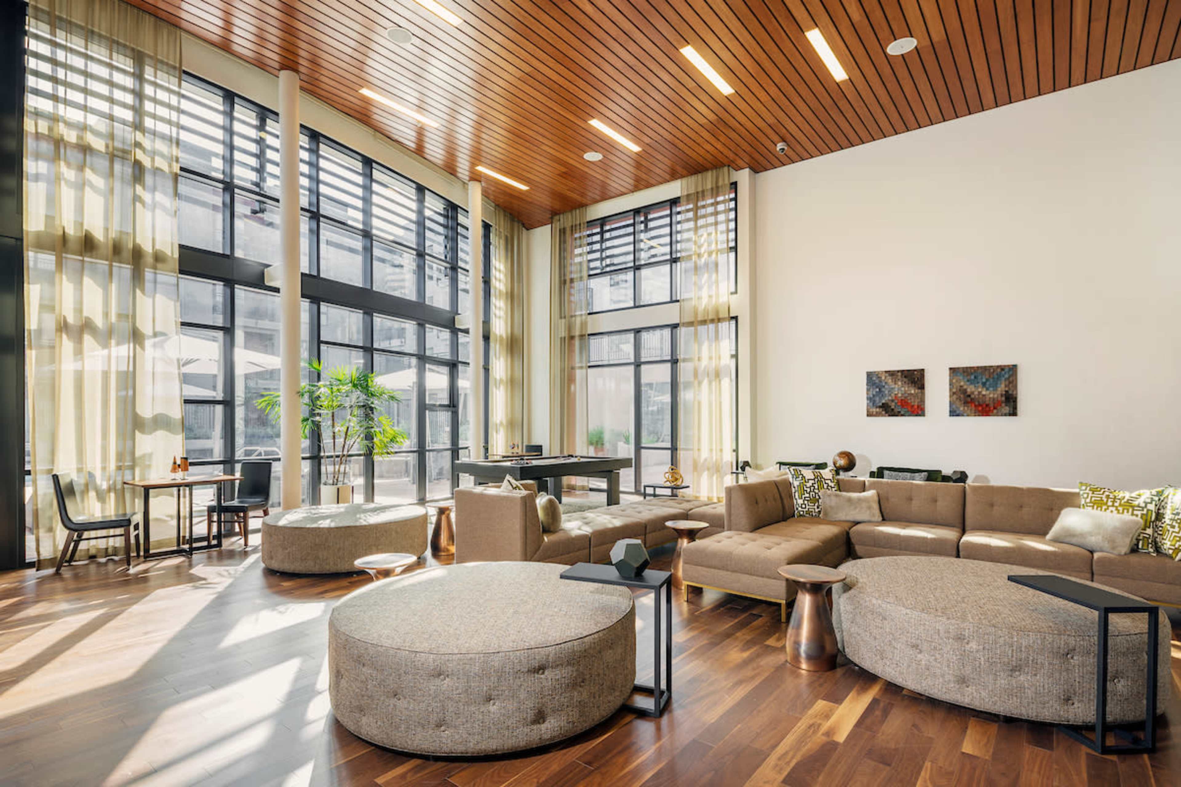 A modern lounge area with large windows, wooden ceiling, and a mix of circular and angular furniture arranged around a central seating space.