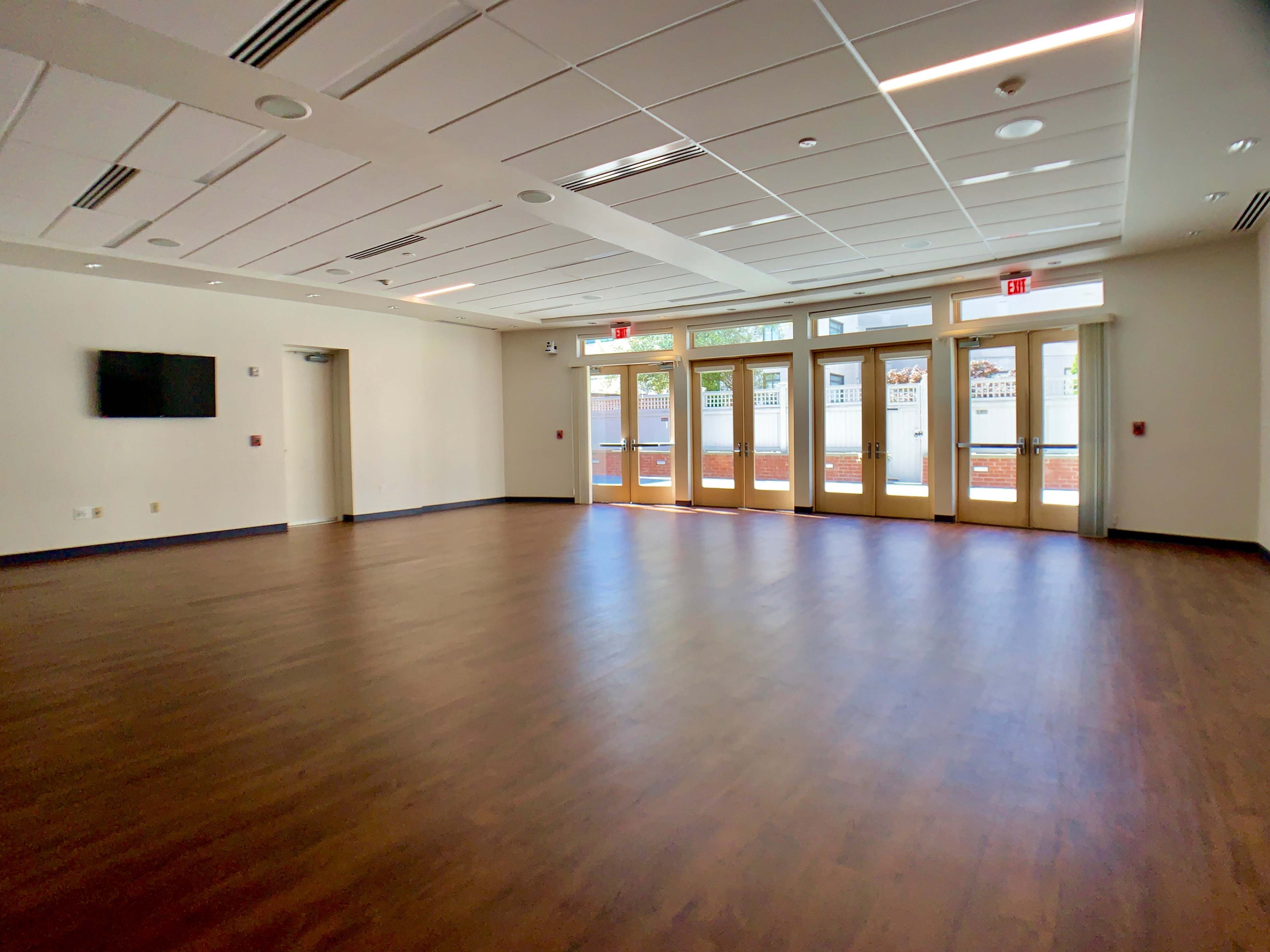 A spacious, empty room with wooden floors, large windows, and double doors that lead outside.
