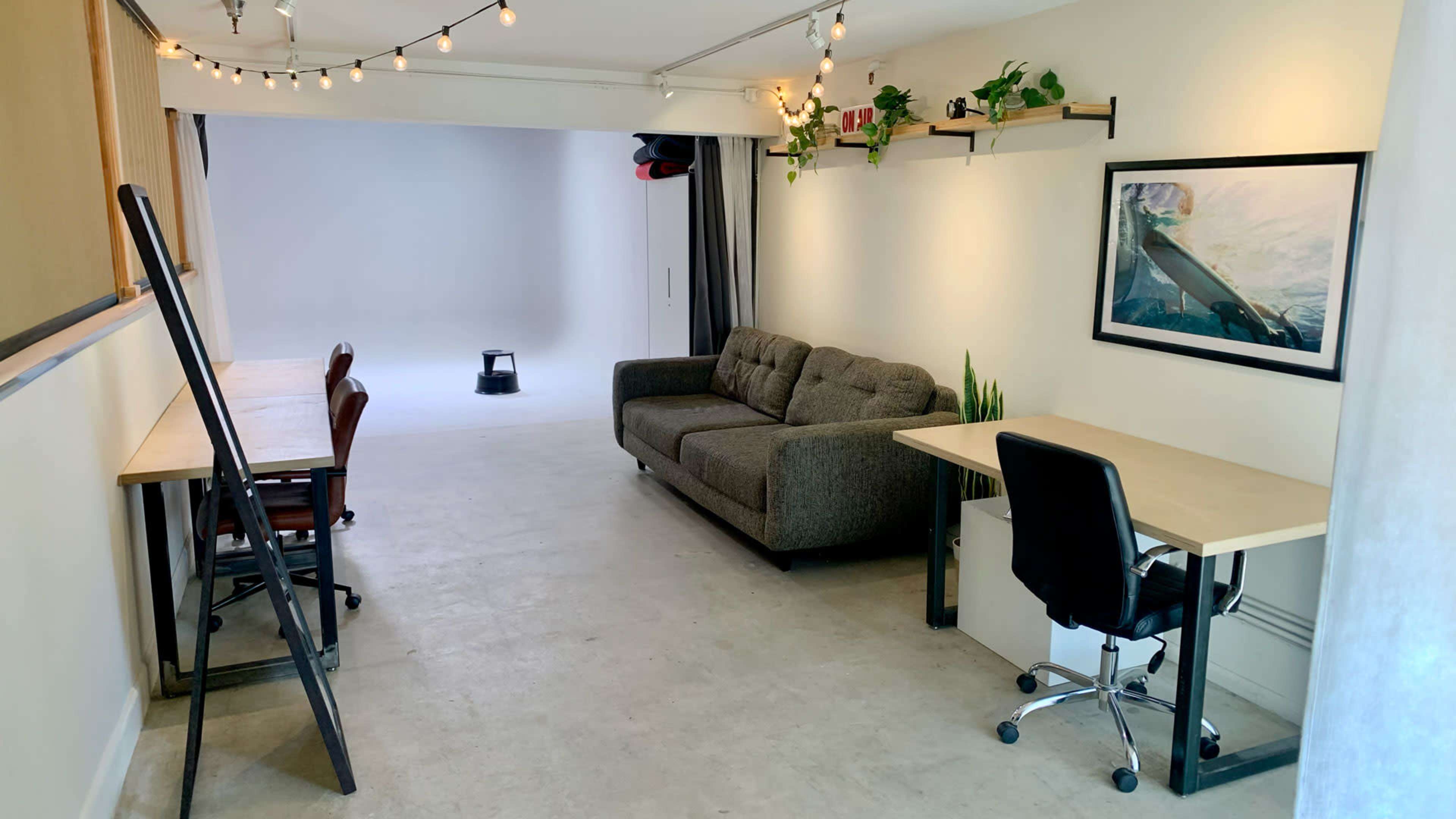 The image shows a minimalist studio space featuring a gray sofa, two wooden desks, plants on shelves, and a light background area.