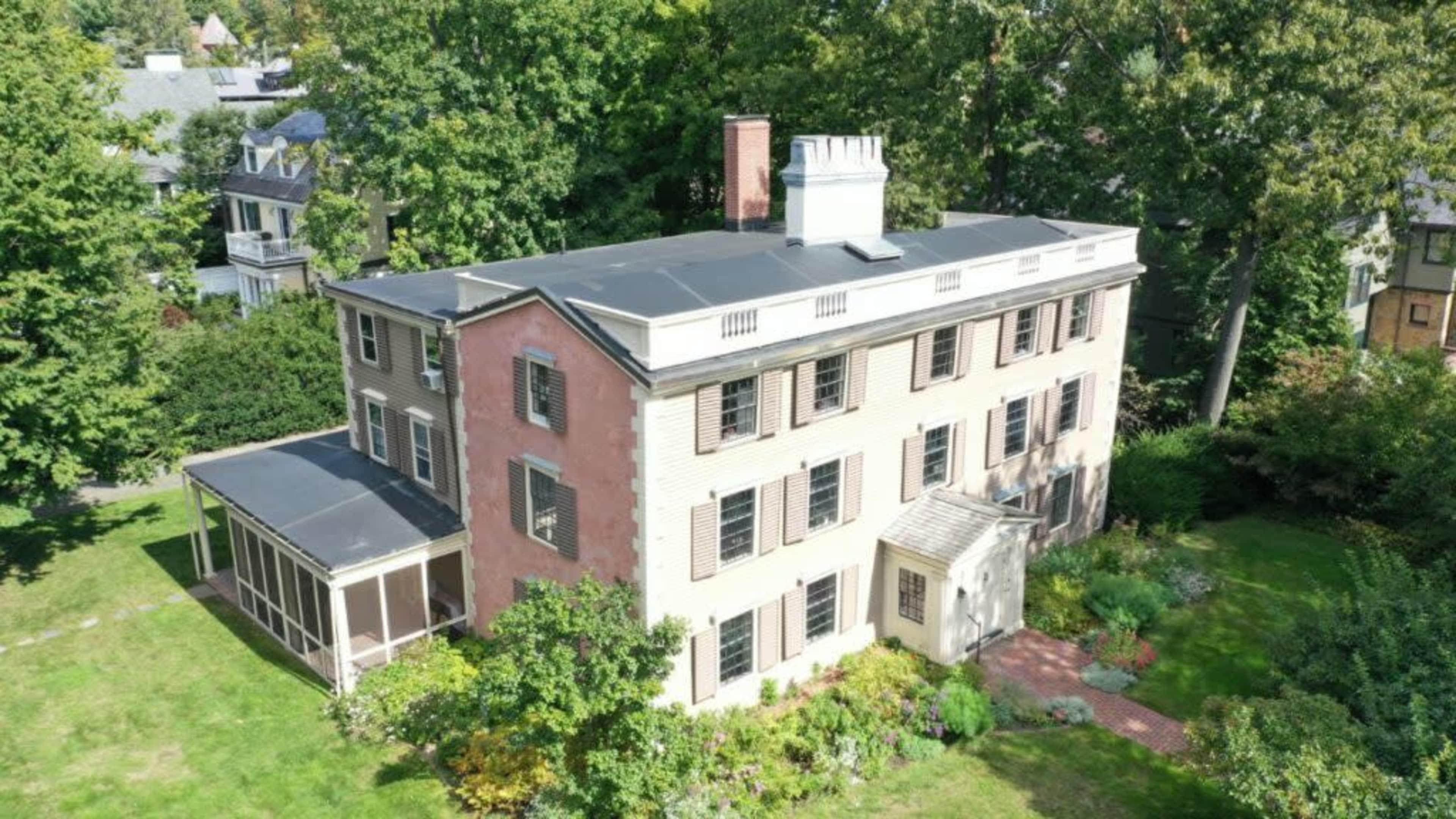 A large, three-story house with a combination of light-colored siding and a screened porch, surrounded by trees and a garden.