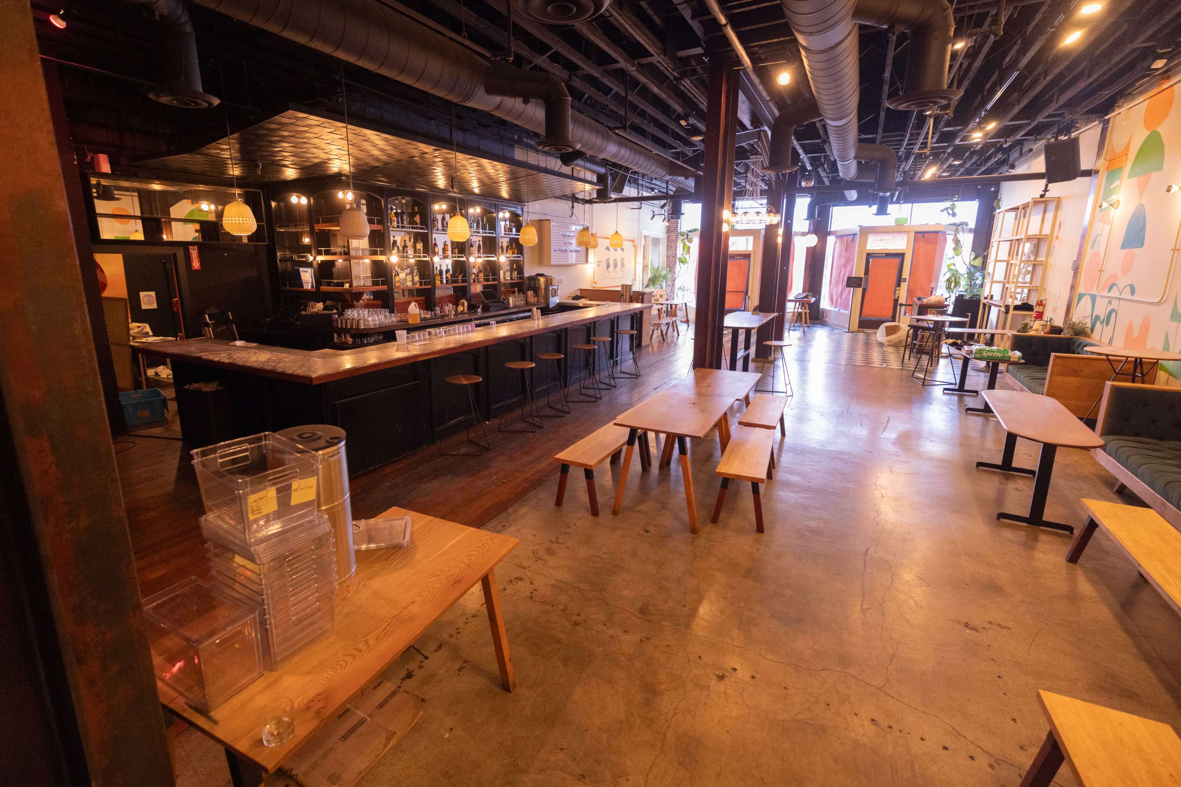 The interior of a spacious café features a long bar area with seating, wooden tables scattered throughout, and large windows that provide natural light.