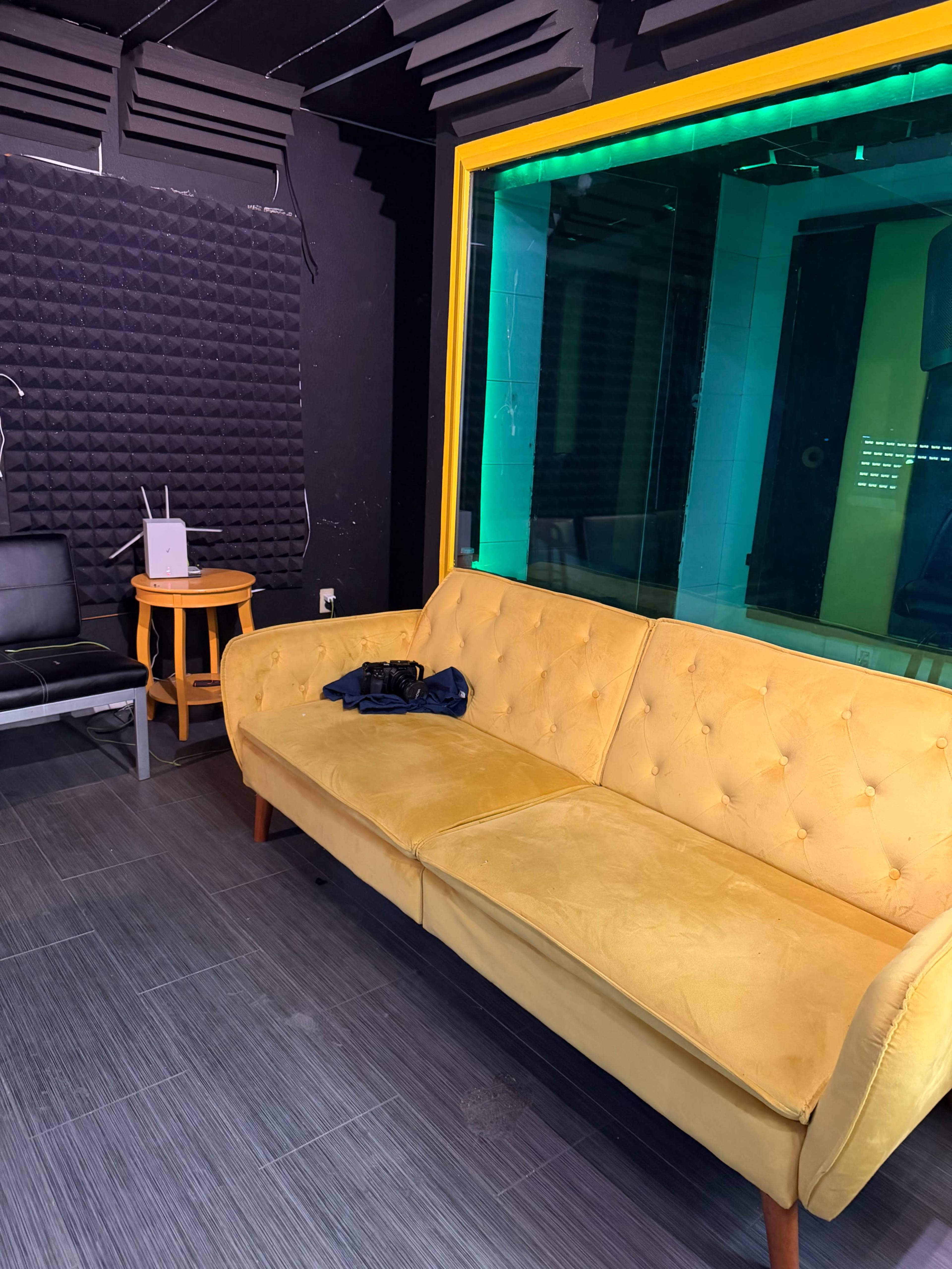 The image shows a yellow couch in a room with dark walls, textured soundproof panels, and a large glass window revealing a sound studio.