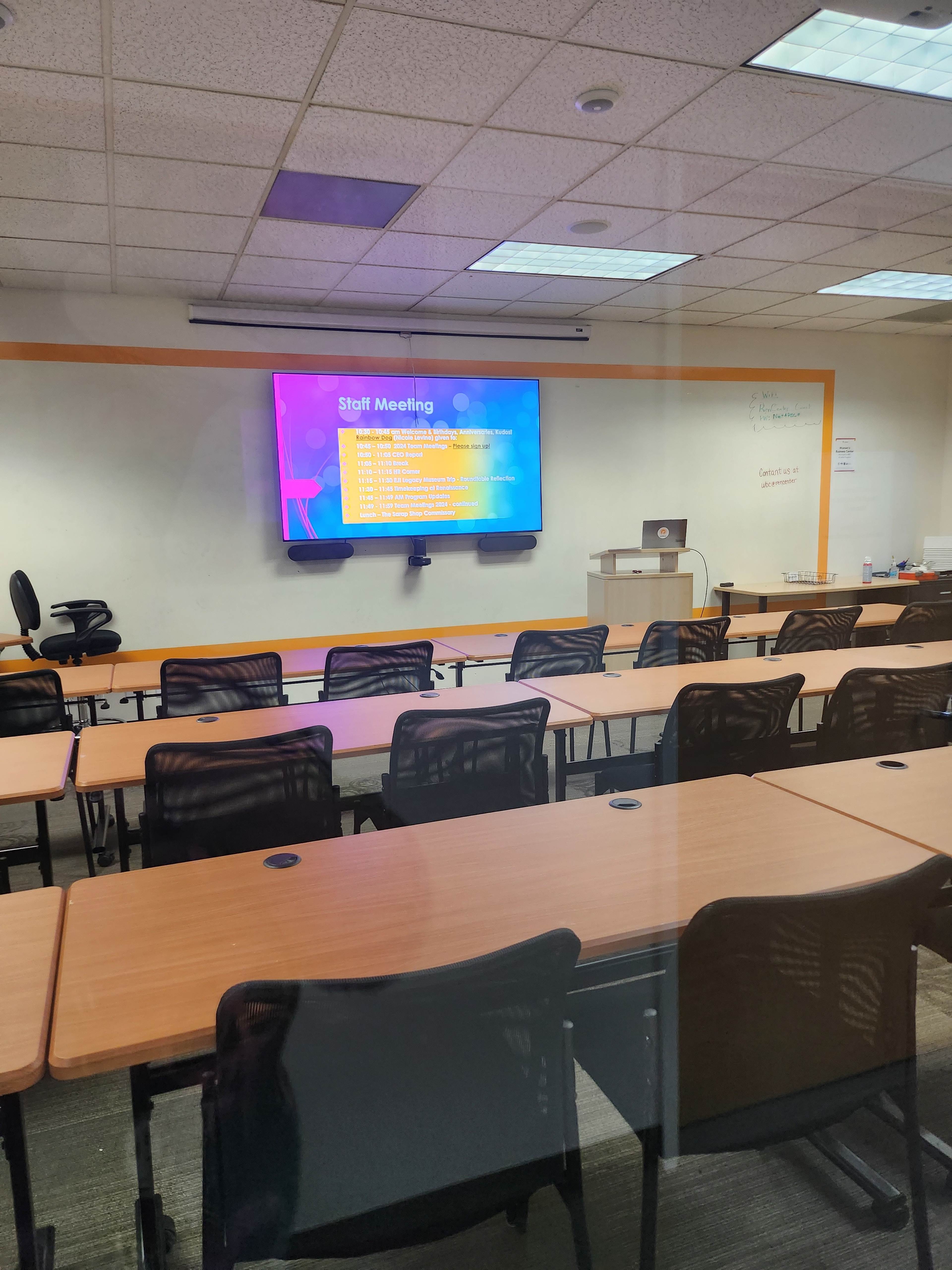 A conference room is set up for a staff meeting, featuring rows of empty chairs facing a screen displaying meeting agenda items.