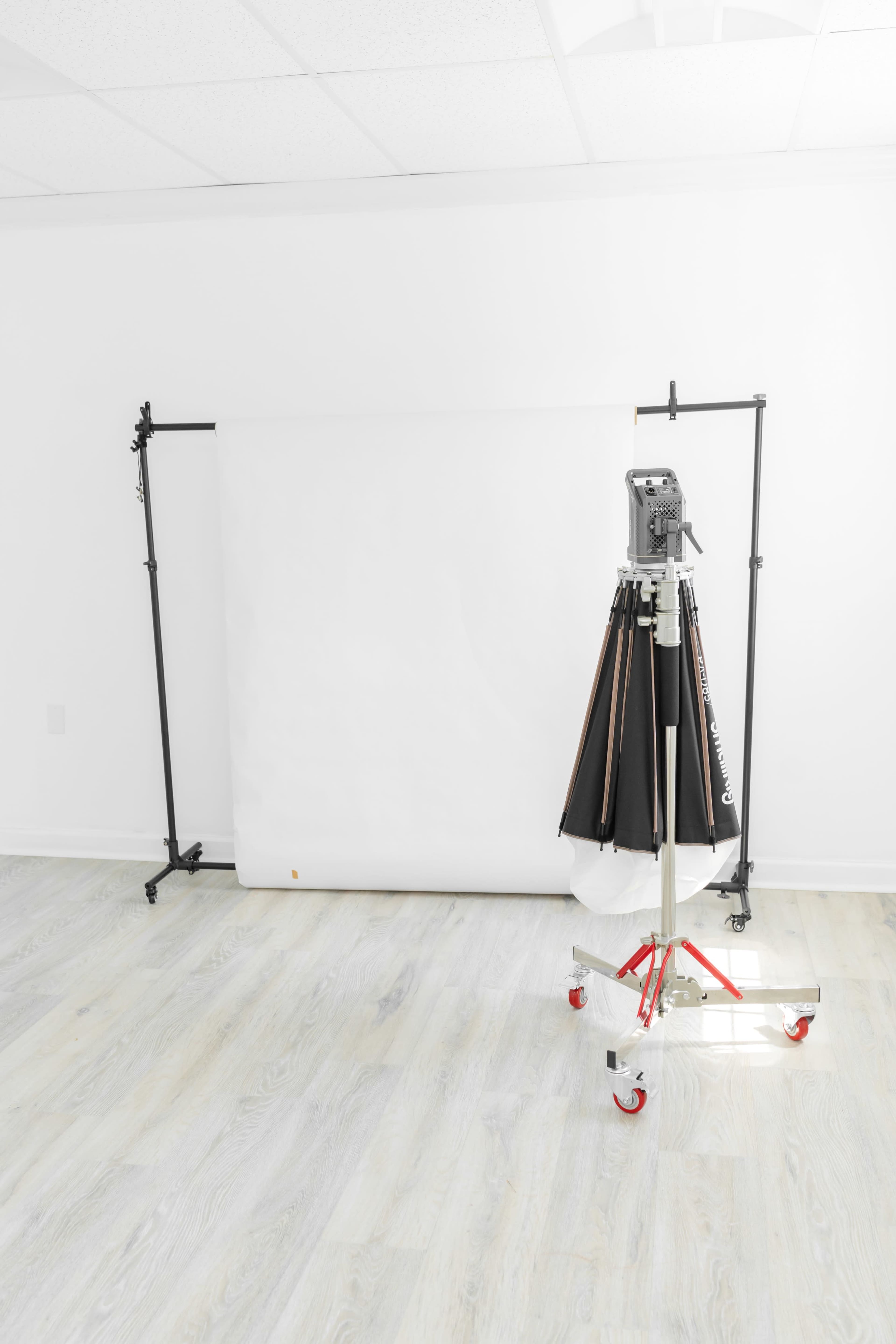 A photography studio setup featuring a white backdrop supported by metal frames and a lighting stand with umbrella reflectors.