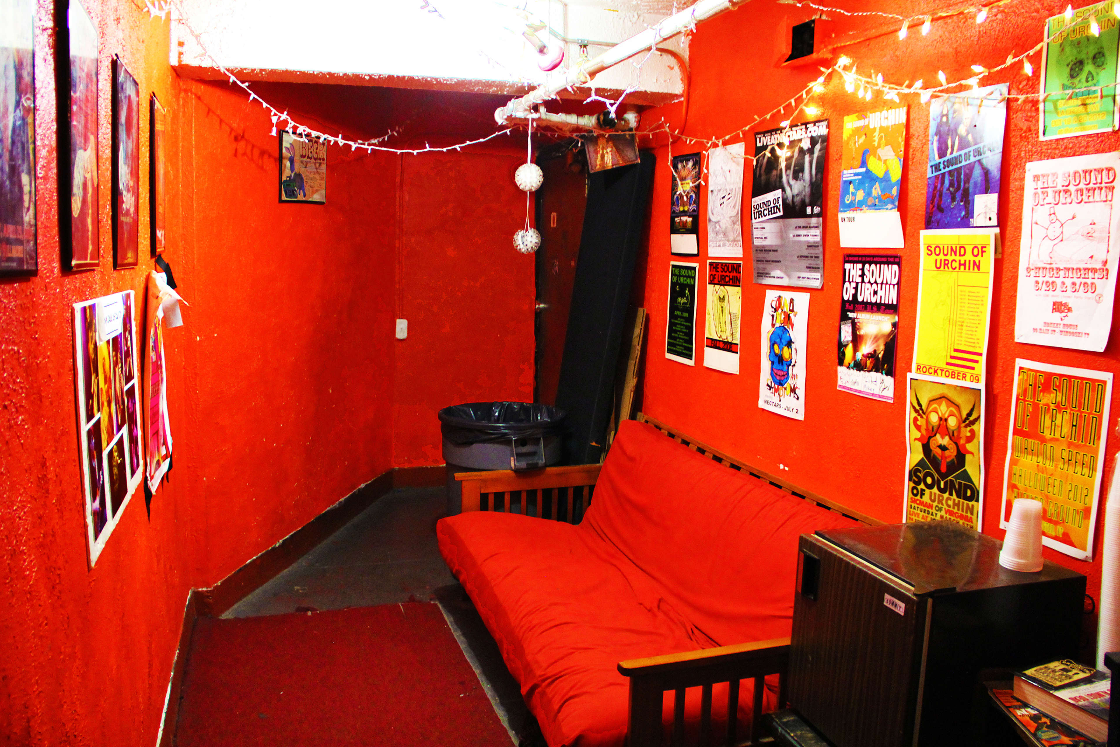 A small, orange-walled room featuring a red couch, a trash can, and various posters on the walls.