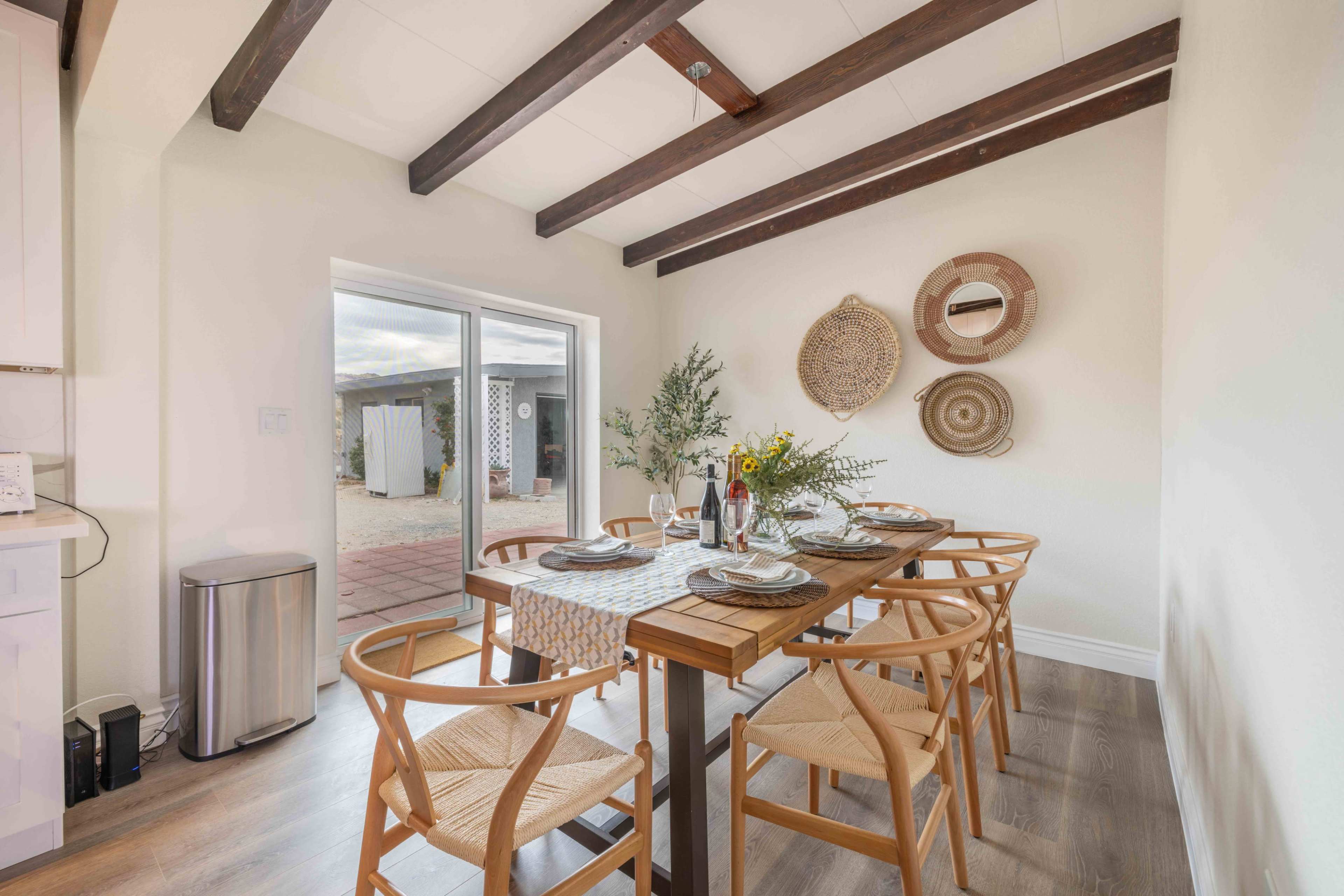 A dining area features a wooden table set for a meal, accompanied by six chairs and decorative wall hangings, with a view of an outdoor patio through sliding glass doors.