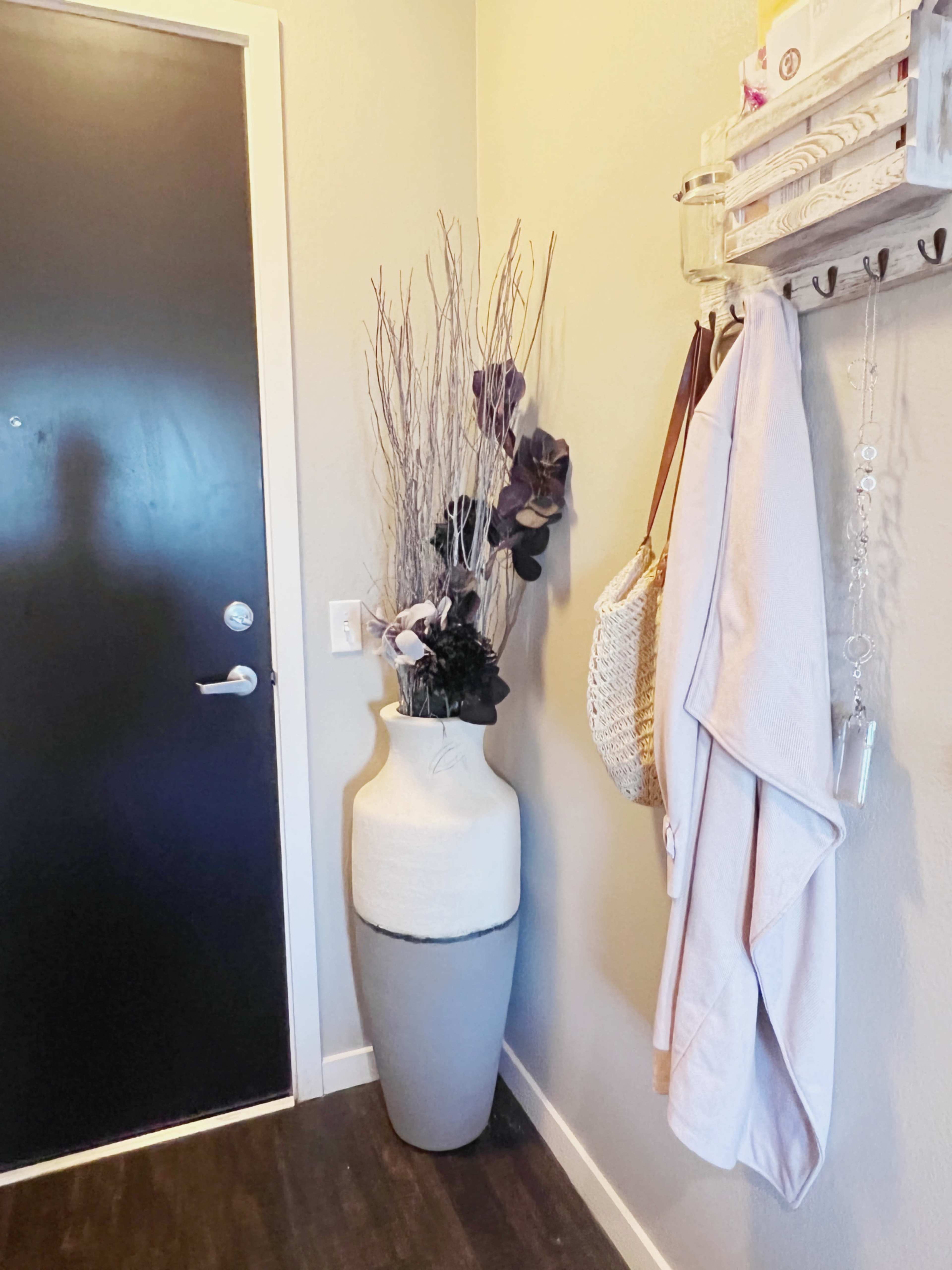 A tall, two-toned vase with branches and flowers stands next to a dark door, while hooks on the wall hold a bag and a towel.